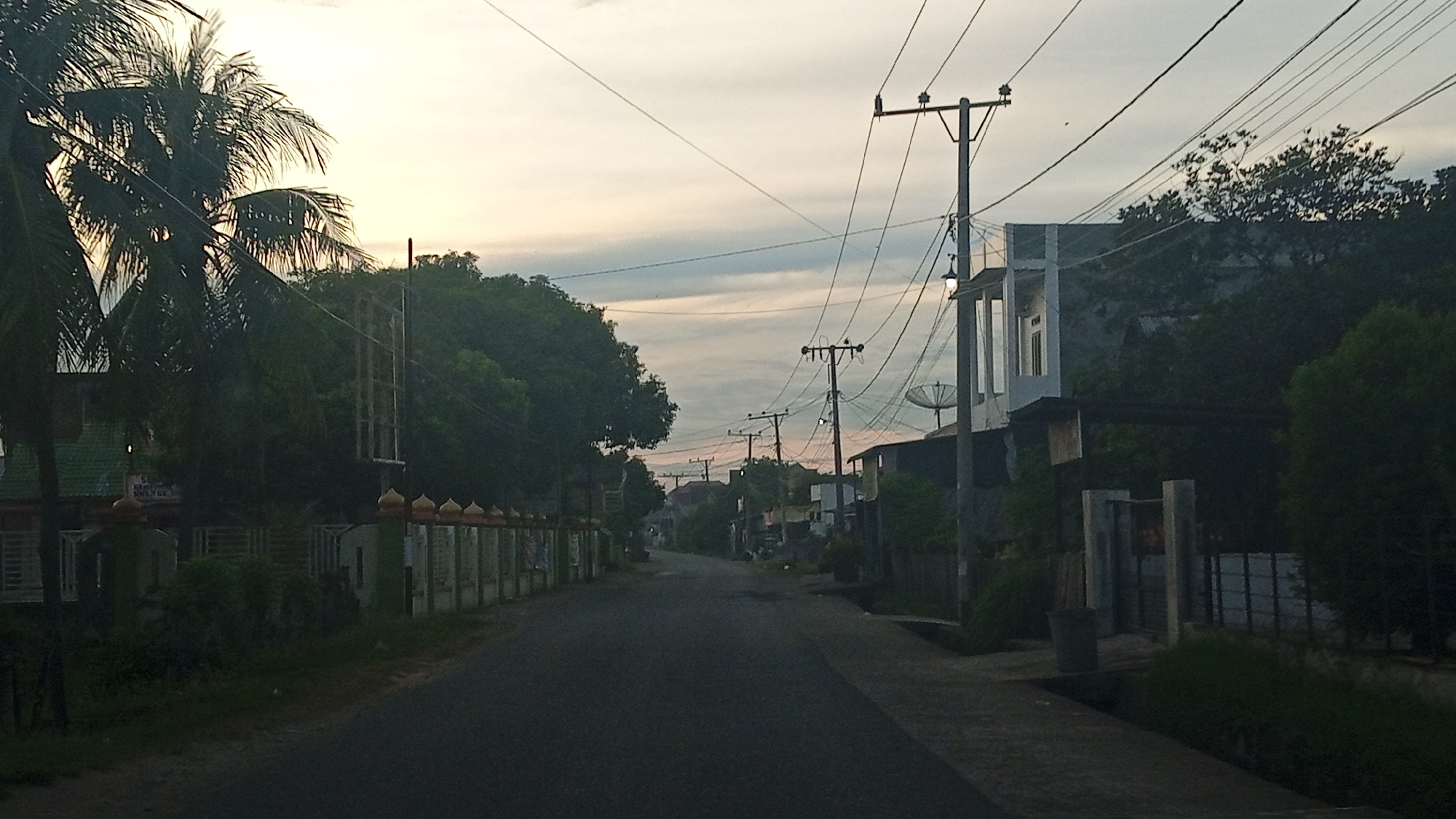 The image depicts a quiet, paved road in Meunasahteungkudigadong, Indonesia, during what appears to be early morning or late evening, indicated by the soft, diffused light and overcast sky. The road is lined on both sides with vegetation, including palm trees on the left and denser trees on the right. Structures are visible along the road, including houses and fences. On the left, a green-roofed building with white fencing and decorative pillars topped with golden domes is present. On the right, a modern, two-story white building with dark trim stands. Numerous utility poles with complex networks of wires crisscross the sky, typical of urban or suburban environments. A satellite dish is visible on the roof of a building on the right. The overall atmosphere is serene and unremarkable, suggesting a typical residential street scene in this Indonesian locale. No people are visible in the frame.