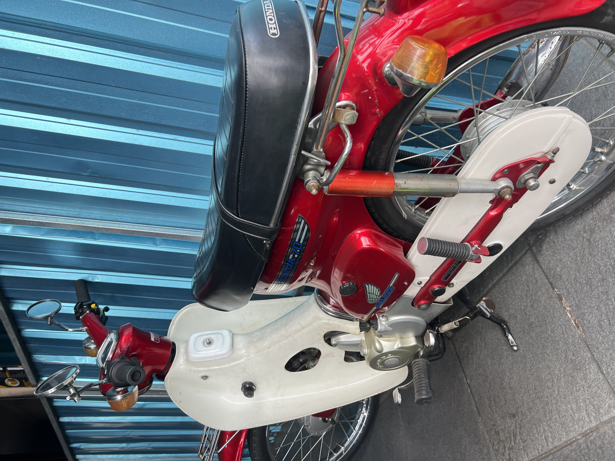 A red and white Honda 70 motorcycle is parked on a tiled floor in front of a blue corrugated metal wall. The motorcycle has a black seat, chrome accents, and a kickstand. The image appears to be taken in the city of Gianyar, Indonesia, given the context of previous images, suggesting it is likely a commercial or residential area within the city. No people are visible in the frame. The lighting suggests it is daytime.