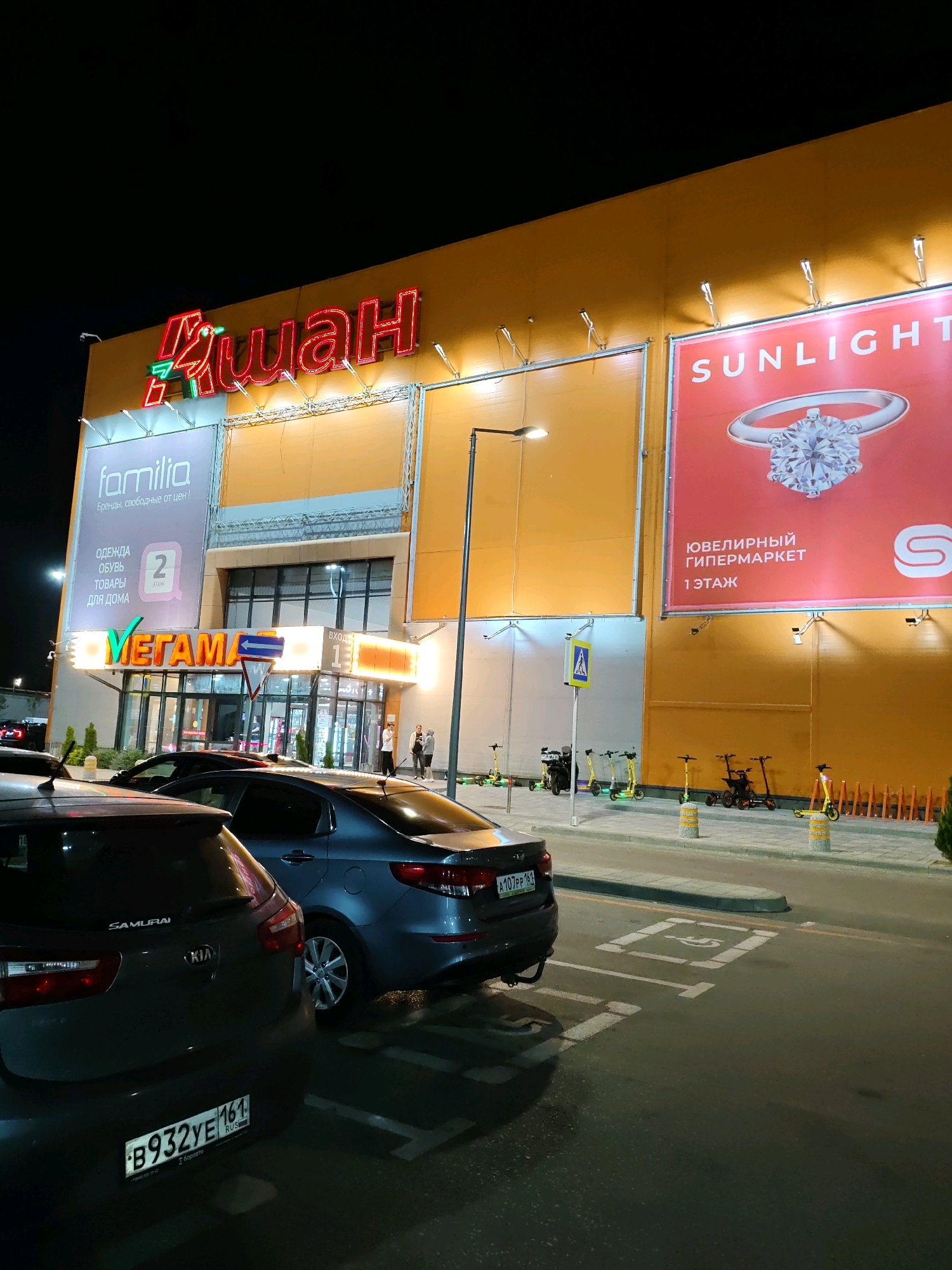 The image shows a night view of the "Auchan" supermarket on Malinovsky Street in Rostov-on-Don, Russia. The bright neon sign of "Auchan" is prominently displayed on the yellow facade of the building. To the right of the main entrance, there is a large advertisement for "Sunlight" jewelry, featuring a diamond ring. Above the entrance, a sign reads "VEGAMA," indicating a shopping area within the supermarket. The entrance itself is illuminated by lights, and people can be seen walking in and out. In front of the building, there is a parking lot with several cars. Electric scooters are parked along the sidewalk. The overall scene is characteristic of a busy retail environment at night.