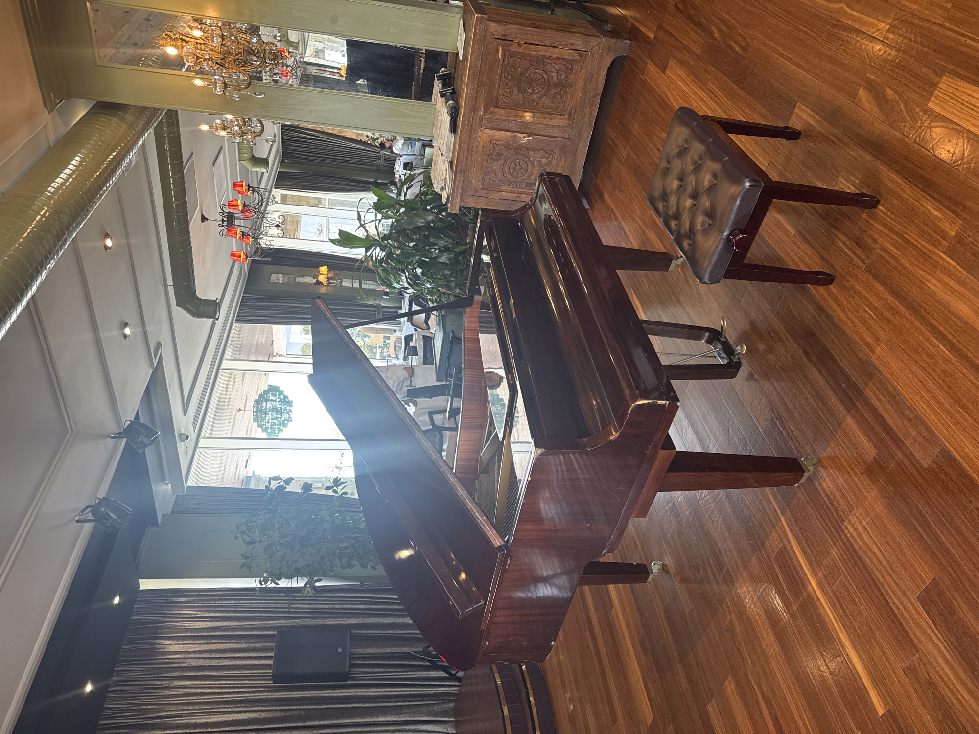 The image depicts a well-lit, upscale indoor setting, possibly a restaurant or event hall, in Sabail Raion, Azerbaijan. A polished, dark wood grand piano dominates the foreground, positioned on a gleaming wooden floor. To the right of the piano is a dark, tufted bench. In the background, through large windows, outdoor elements are visible. The interior features decorative lighting, including chandeliers and track lighting, and large, dark curtains. Green foliage is present, adding a touch of nature to the environment. A vintage wooden cabinet with ornate carvings stands to the right of the piano, and a speaker is visible to the left. Several people are seated at tables in the background, suggesting a social gathering or dining event. The overall impression is one of elegance and sophistication.
