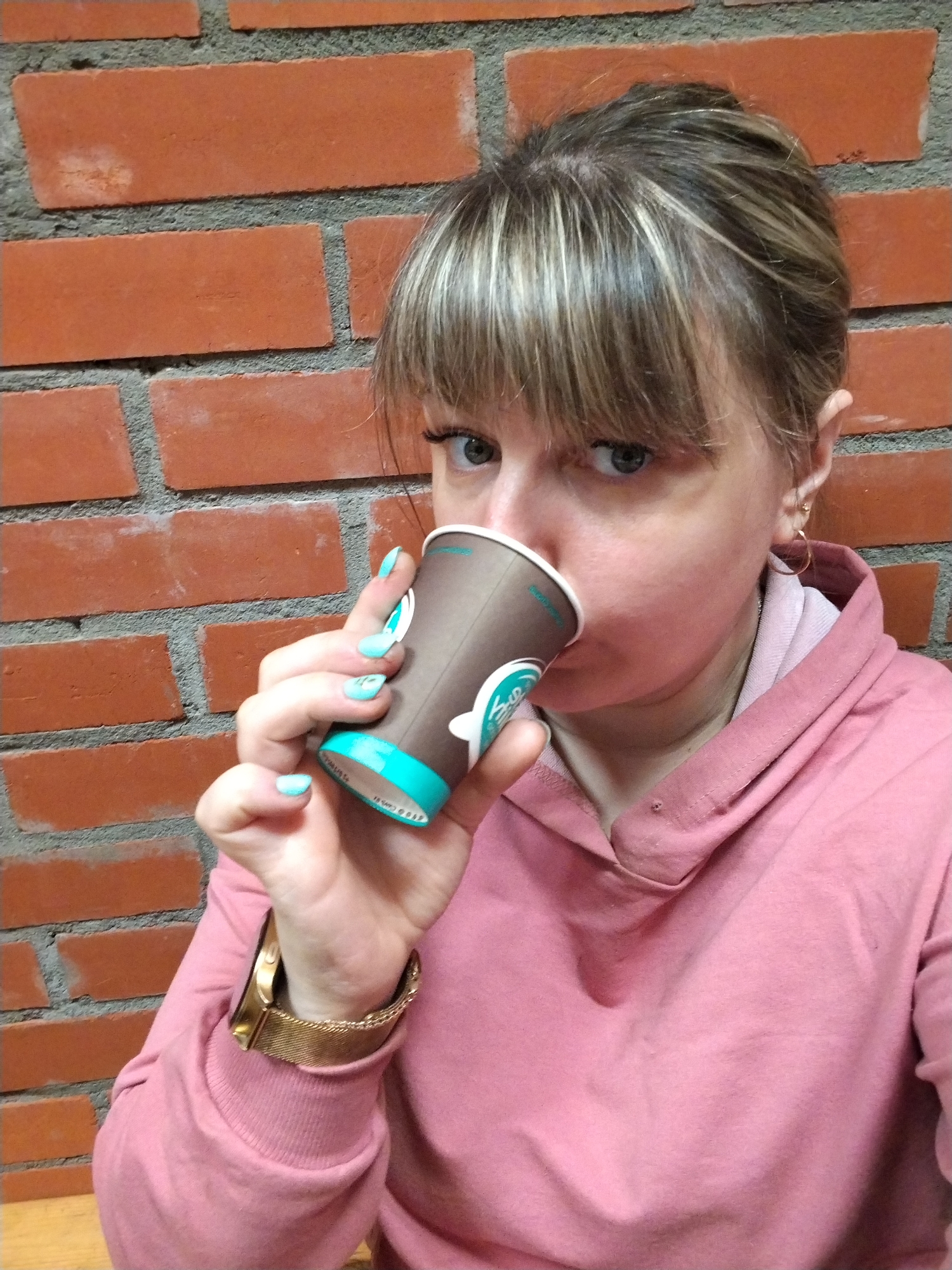 An eye-level portrait captures a woman drinking from a paper coffee cup against a distinct red brick wall, likely in Шебунино, Russia. The woman has light brown hair with blonde highlights, styled with bangs across her forehead, and her blue-grey eyes engage directly with the viewer. She is dressed in a soft pink hooded sweatshirt. Her left hand, adorned with light teal nail polish, holds the brown paper cup to her lips. On her left wrist, a gold-toned watch with a mesh band is visible, and a small gold hoop earring is in her right ear. The coffee cup features a light teal bottom rim and a partially visible logo with text. Though mostly illegible, fragments like 'Canals' or 'Canal's' and a copyright notice '© ALL RIGHTS RESE' can be discerned. The background consists of reddish-orange bricks with prominent grey mortar lines. Her expression is calm and direct, suggesting a candid moment of pause.