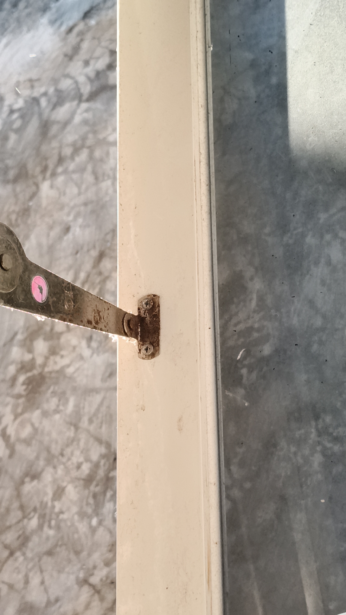 The image shows a close-up view of a light-colored door frame with a rusty metal hinge attached. The hinge is secured to the frame by two screws. A small pink circular sticker is visible on the hinge. The background consists of a textured, grey surface, possibly concrete or a similar material. The scene appears to be outdoors, with a shadow cast from the right side of the frame. The overall impression is one of a functional detail of a door, possibly in an urban or industrial setting. There are no people or discernible actions occurring in the image. The location in Kerep Kidul, Indonesia, is not visually evident from the image itself.