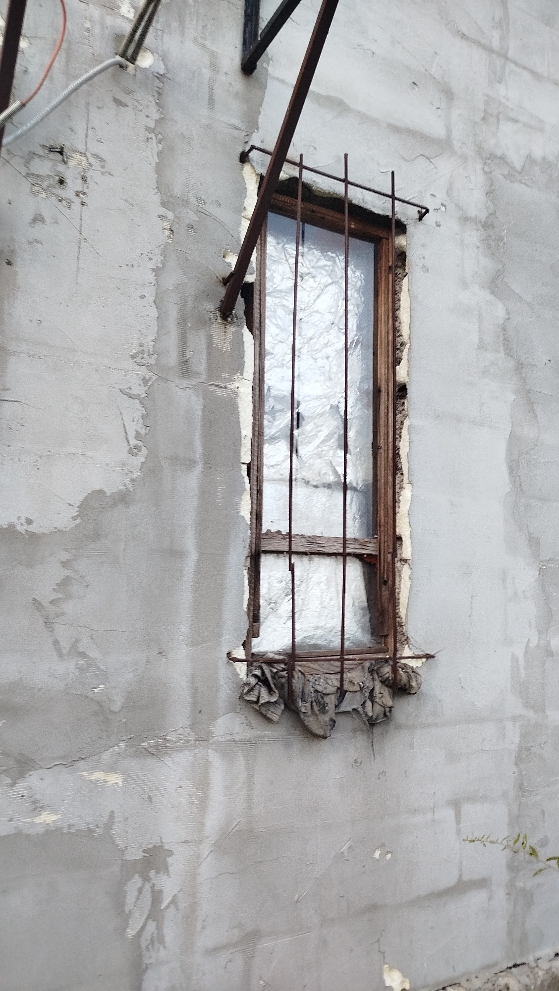 The image shows a close-up of a dilapidated window on a building's exterior wall. The wall is made of grey concrete with visible cracks and patches of older plaster underneath. Some of the plaster has chipped away, revealing a textured underlayer. A metal pipe runs diagonally across the upper left portion of the wall, attached to a wire conduit.
The window itself is recessed into the wall, with a wooden frame that appears weathered and damaged. The glass is covered with crumpled, translucent plastic sheeting, obscuring any view inside. Horizontal metal bars are bolted to the exterior of the window frame, serving as a security grate. Below the window, at the bottom of the frame, is a pile of dark, tattered fabric, likely intended to block drafts.
The overall scene suggests neglect and disrepair, consistent with an abandoned or derelict structure. This window is located on a building in the city of Kostiantynivka, Ukraine.