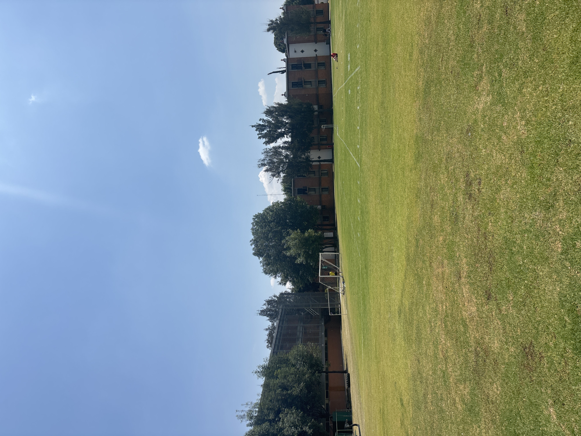 The photograph captures a daytime scene at a sports field in San Andrés Cholula, Mexico. The field, primarily green grass, is lined and appears to be used for football. A goal post is visible near the middle of the image. In the background, a multi-story brick building is partially obscured by trees. The sky is blue with a few clouds, and the sun is shining. In the distance, a person wearing red clothing is located near the field markings. The overall environment is outdoors with the intersection of sports, nature, and architecture.
