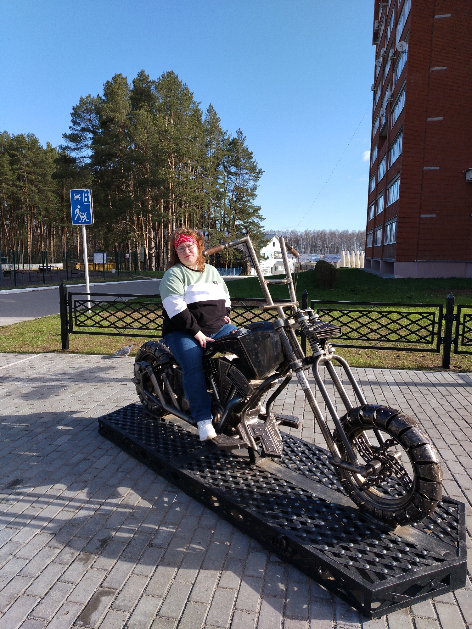 A young woman with red hair sits on a metal sculpture of a motorcycle in Chaikovsky, Russia. She wears a red bandana, glasses, a green, white, and black sweatshirt, blue jeans, and white shoes. The motorcycle sculpture is made of metal and sits on a raised platform of black metal mesh. Behind her is a black metal fence, a pedestrian and bus stop sign, a row of tall pine trees, and a brick building. The ground is paved with grey bricks. The sky is blue with some light clouds. The scene is well-lit.