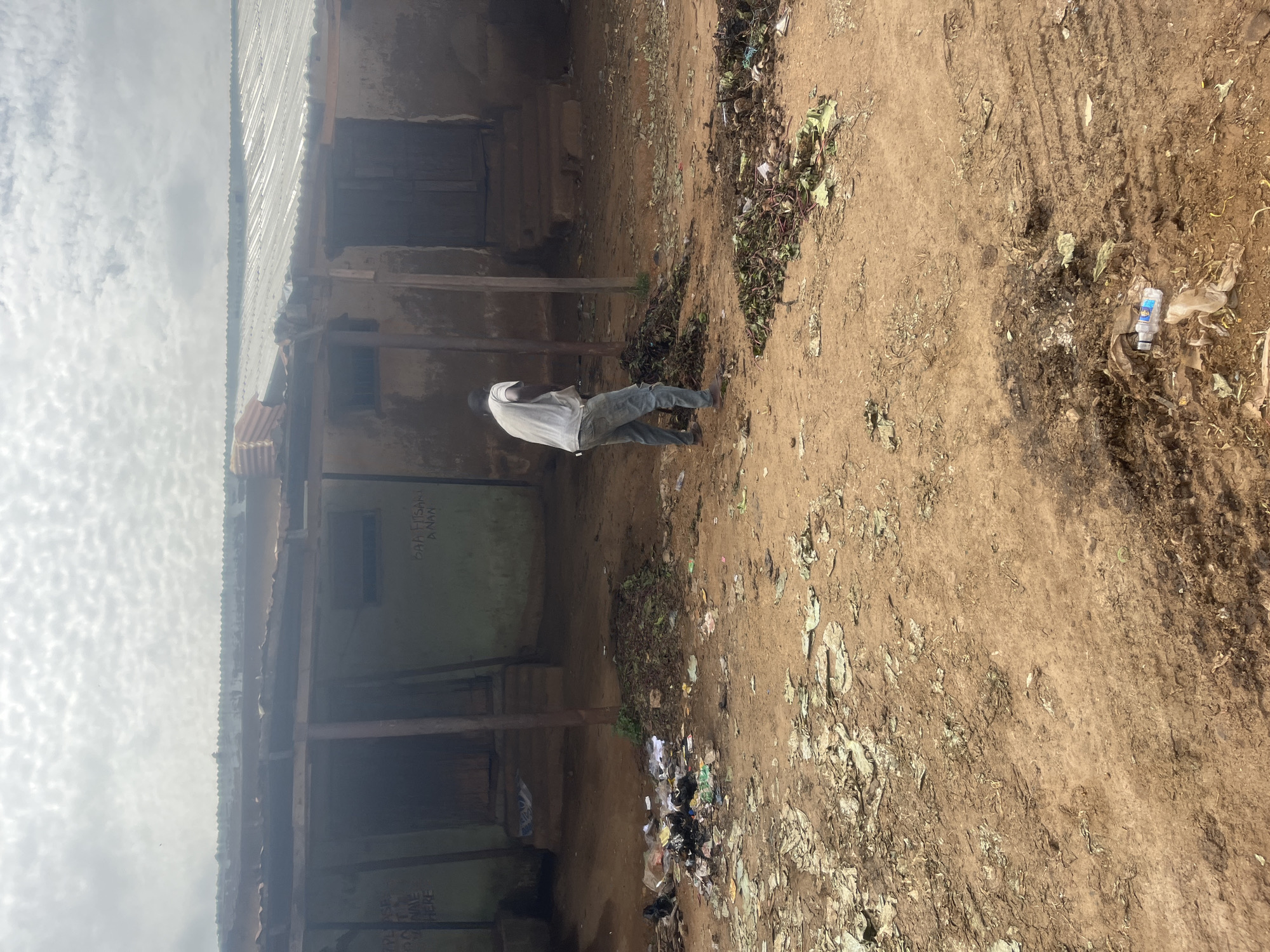 A man in a white shirt and blue jeans stands in the yard of a dilapidated building in Gura Top, Nigeria. The yard is littered with trash and debris. The building has a corrugated metal roof and wooden posts supporting the structure. Graffiti is visible on the building's walls. A plastic water bottle is on the ground. The sky is overcast with clouds.