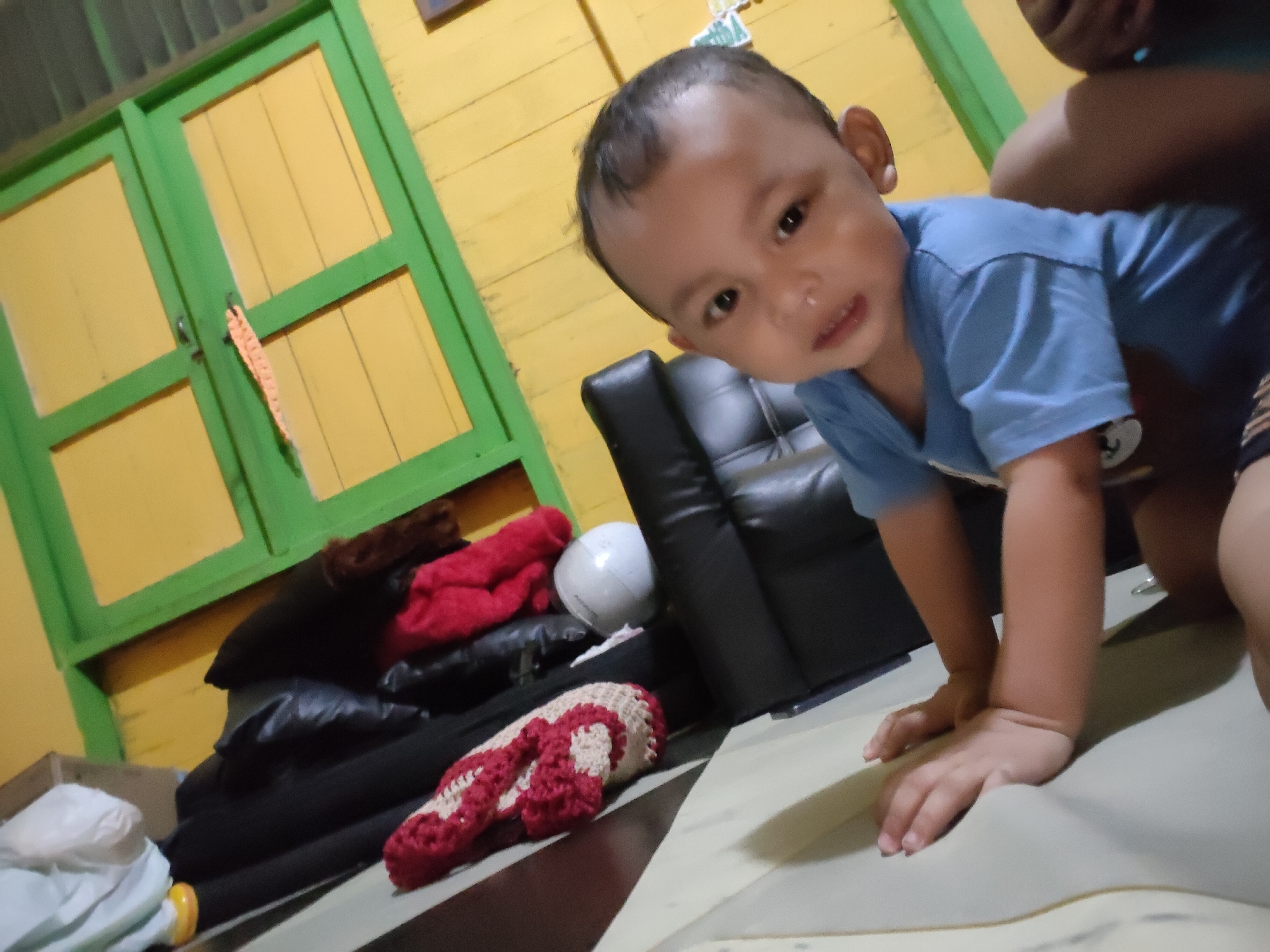 A close-up, low-angle shot captures a young child wearing a blue t-shirt, positioned on a light-colored surface in what appears to be a residential setting. The child is leaning forward, with their hands resting on the surface, looking towards the camera with a neutral expression. In the background, a yellow wall with green trim framing two closed doors is visible. To the right of the child, a black leather armchair and a pile of folded red and black fabric items are present. The overall impression is of an informal, indoor environment, possibly a living room or play area. The specific location in Tanah Grogot, Indonesia, is not discernible from the visual elements of the image.