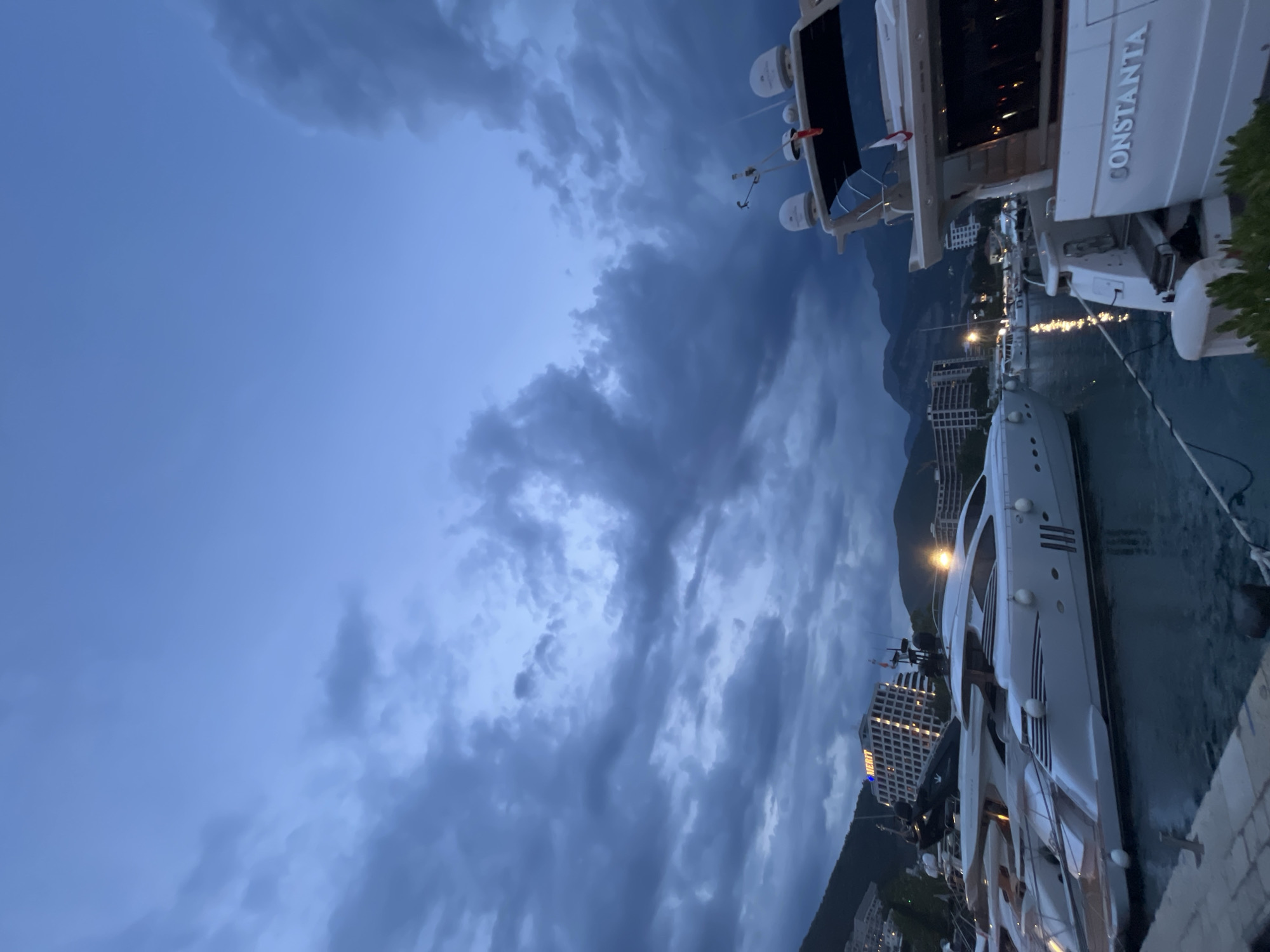 The image shows a nighttime view of the Budva Marina in Budva, Montenegro. The sky is dark blue with scattered clouds, and lights from buildings and boats illuminate the scene. Several yachts are docked in the marina, with their sleek designs visible in the low light. In the background, buildings and mountains are silhouetted against the night sky. One yacht in the foreground has "CONSTANTA" written on its stern. The overall impression is one of a tranquil, upscale waterfront at dusk or night.