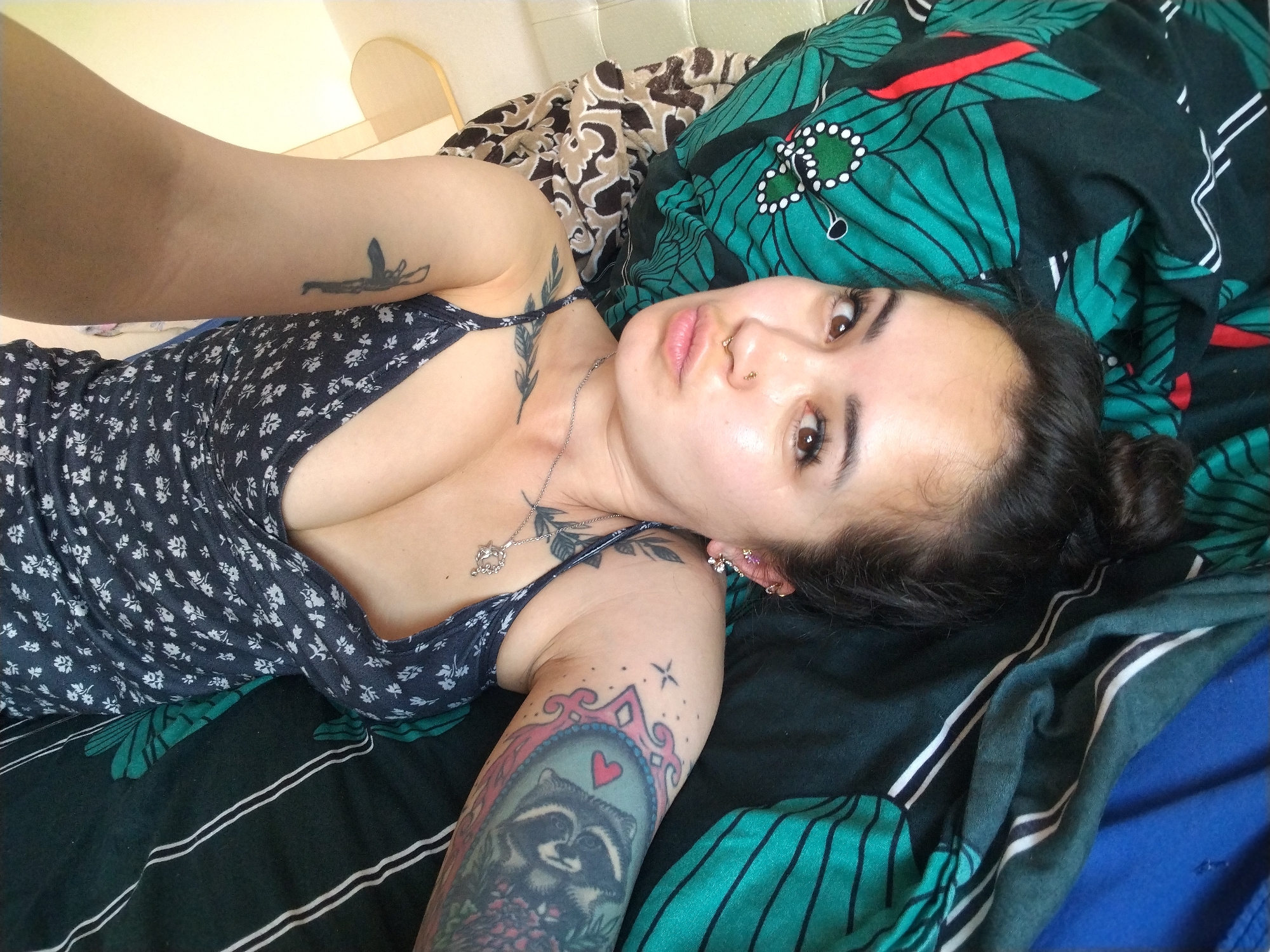 A young woman with dark hair pulled up and dark brown eyes is lying on her side in a bed. She is wearing a dark blue floral print dress and has several tattoos visible on her arms and chest. One tattoo on her upper arm depicts a raccoon within a heart-shaped frame, surrounded by flowers. She is wearing earrings and a nose ring. The bed is covered with patterned bedding, including a green, teal, and red floral print and a blue blanket. The background is blurry but appears to be a room with a headboard. The image is a close-up, selfie-style shot taken from a high angle. The location is identified as Noviy, Russia.