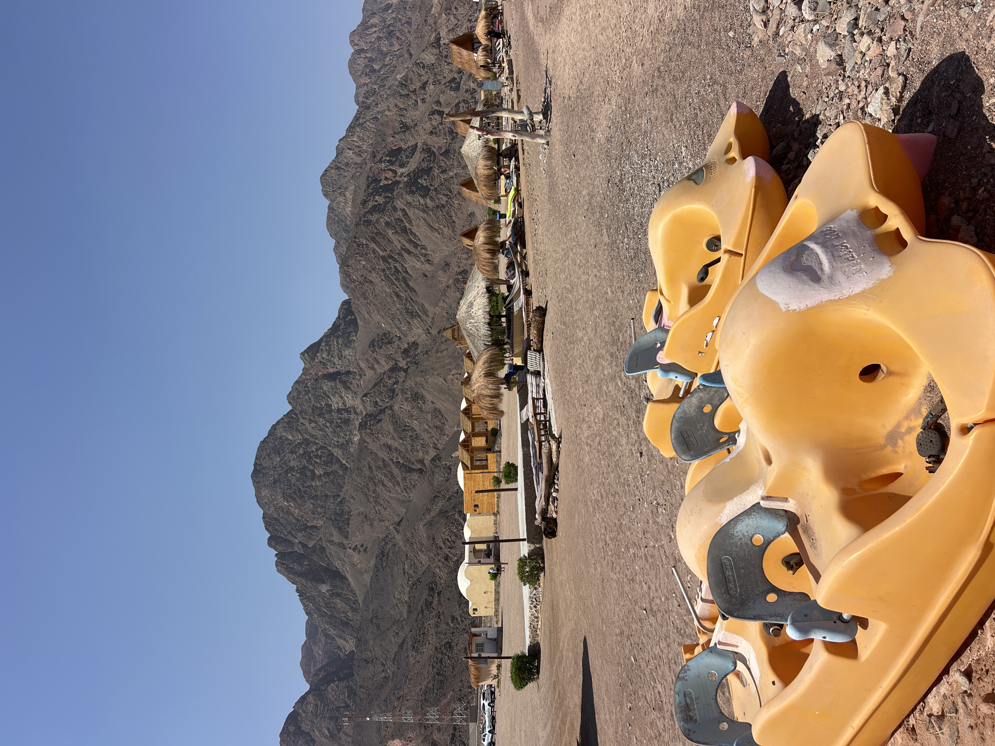 In an outdoor scene in Unknown, Egypt, yellow paddle boats are stacked on a gravelly beach. In the foreground, two paddle boats are partially visible, with their hulls resting on the ground. Their brightly colored surfaces are marked with wear and tear. Behind the boats, a strip of beach is lined with straw-roofed huts and lounge chairs, suggesting a resort or recreational area. Beyond the huts, a range of rugged, barren mountains rise against a clear blue sky. The landscape appears dry and arid, characteristic of a desert environment.