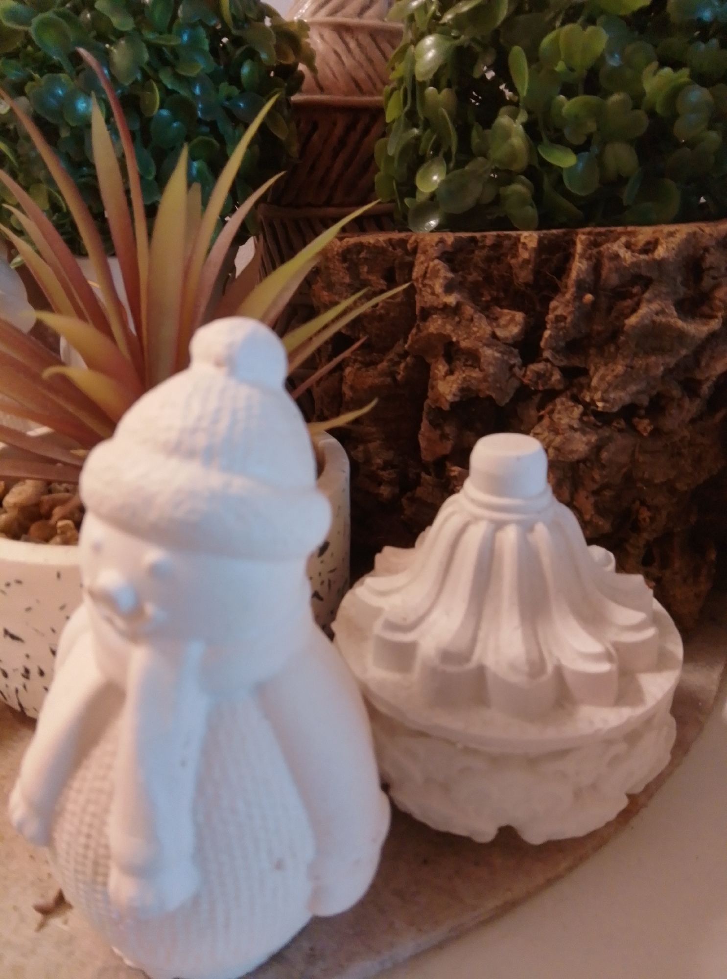 The image depicts a still life arrangement with two unpainted plaster figurines. In the foreground on the left is a snowman figurine wearing a hat and scarf. To its right is a lidded jar or pot with a decorative lid. Behind the figurines, a plant in a speckled pot is visible, along with a textured, bark-like decorative element and foliage of a potted plant. The scene is set on a surface that appears to be a table or shelf. The overall impression is of items prepared for painting or crafting. The image was taken indoors, likely in a home or studio environment. There is no information available to place this image in Wedel, Germany.