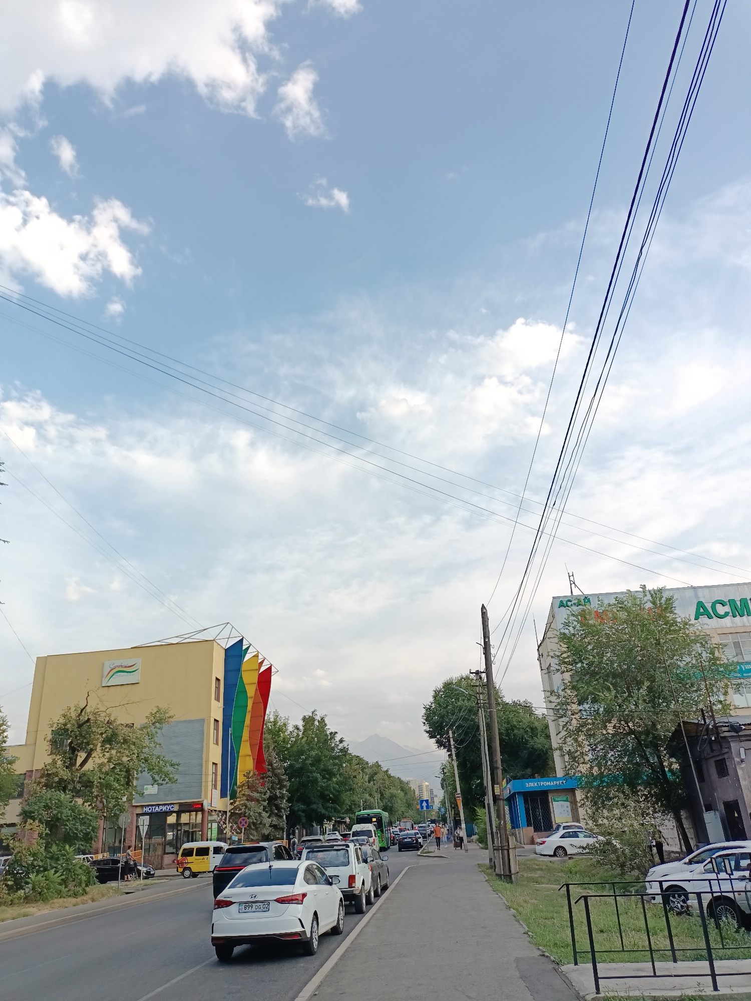 The image shows a street scene in Almaty, Kazakhstan, with a focus on the architecture and traffic. The street is lined with buildings on both sides. On the left, a yellow building with a sign that appears to say "нотариус" (notary) is visible, adorned with colorful vertical banners resembling a rainbow. On the right, a white building with green lettering that reads "АСМ" is prominent, with trees in front of it. The street is busy with various cars, including a white Hyundai, a white SUV, and a green bus. Pedestrians are seen walking on the sidewalk. Overhead, power lines stretch across the frame, and the sky is partly cloudy. In the distance, mountains are faintly visible, indicating the city's geographical setting. The overall impression is a typical urban environment with signs of daily life and commerce.