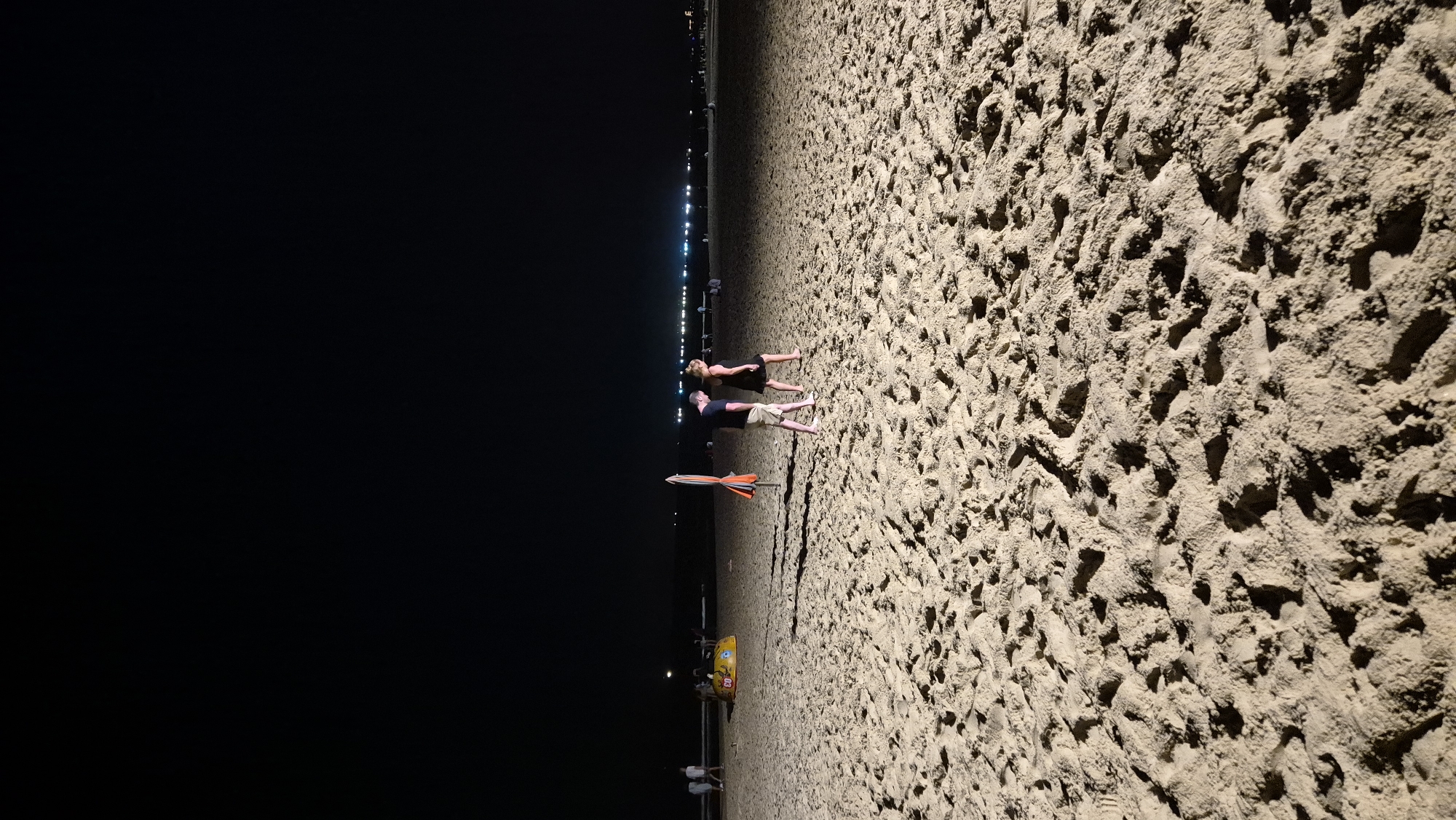 At night on a sandy beach in Da Nang, Vietnam, two individuals, a man and a woman, walk side-by-side. The man, wearing a dark t-shirt and light-colored shorts, walks slightly ahead of the woman, who is dressed in a dark dress. They are positioned in the mid-ground of the image. Further back on the beach, a closed orange and white umbrella stands vertically. To the left and further in the distance, a yellow inflatable object, possibly a ride or toy, is visible. The dark expanse of the night sky stretches above, while the foreground is dominated by the texture of the sandy beach, marked by footprints and shadows cast by unseen lighting. In the far distance, a line of lights suggests the presence of buildings or a developed area along the coastline.