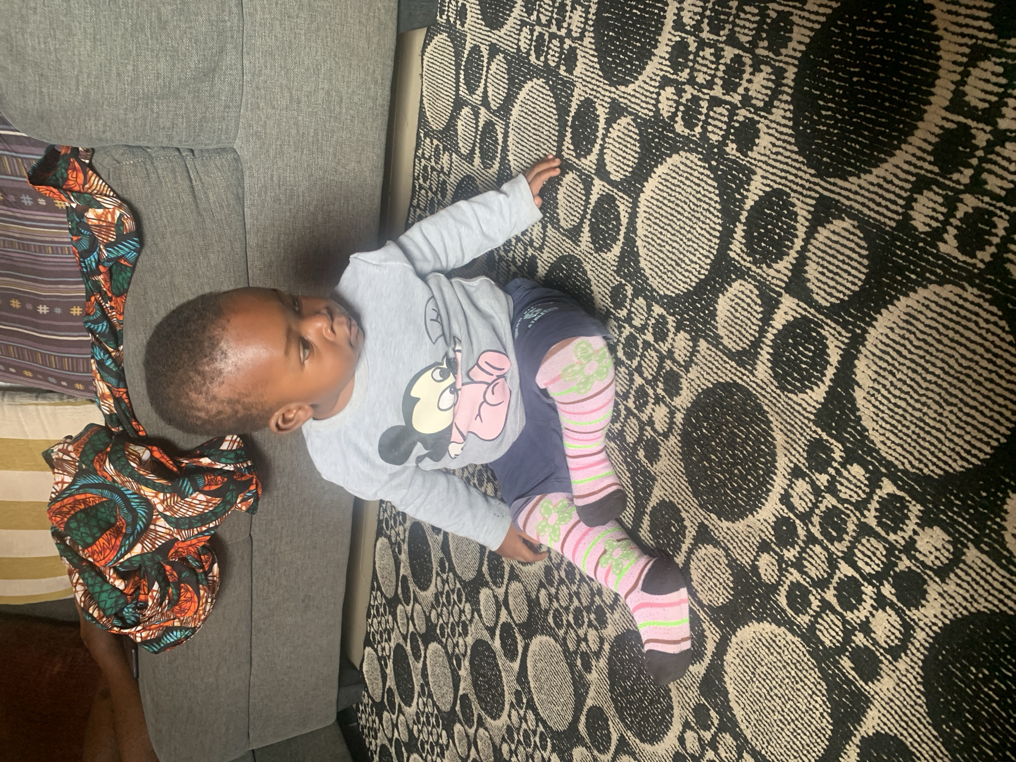 An African child sits cross-legged on a patterned rug in a living room in Sabon Tasha, Nigeria. The child, wearing a grey long-sleeved shirt with Mickey Mouse printed on it, dark blue shorts, and pink and green striped socks, looks down at their hands. A grey sofa with a colorful African print fabric draped over it is visible behind the child. The rug features a repeating circular and geometric pattern in black and beige. The lighting suggests it is daytime, and the overall scene is domestic.