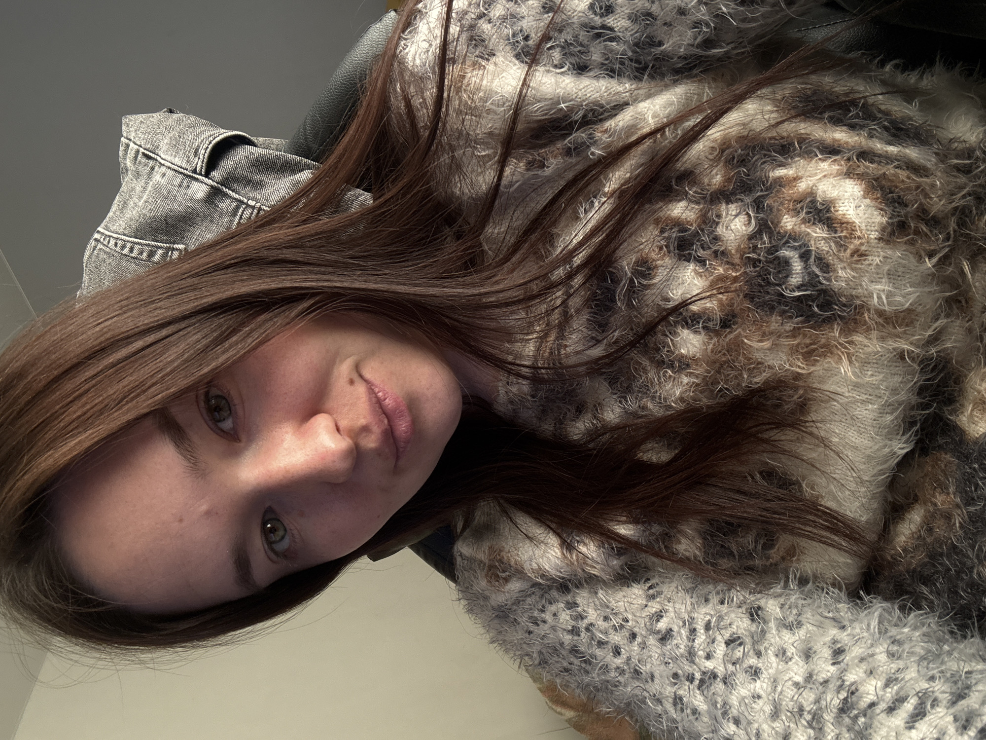 A woman with brown hair and fair skin is positioned at an angle, facing slightly to the left. She is wearing a fluffy sweater with a pattern of gray, white, brown, and black. Behind her, there is a gray denim jacket and a black leather chair. The background appears to be a plain wall. There is no discernible information to establish the location as Фоки, Russia.