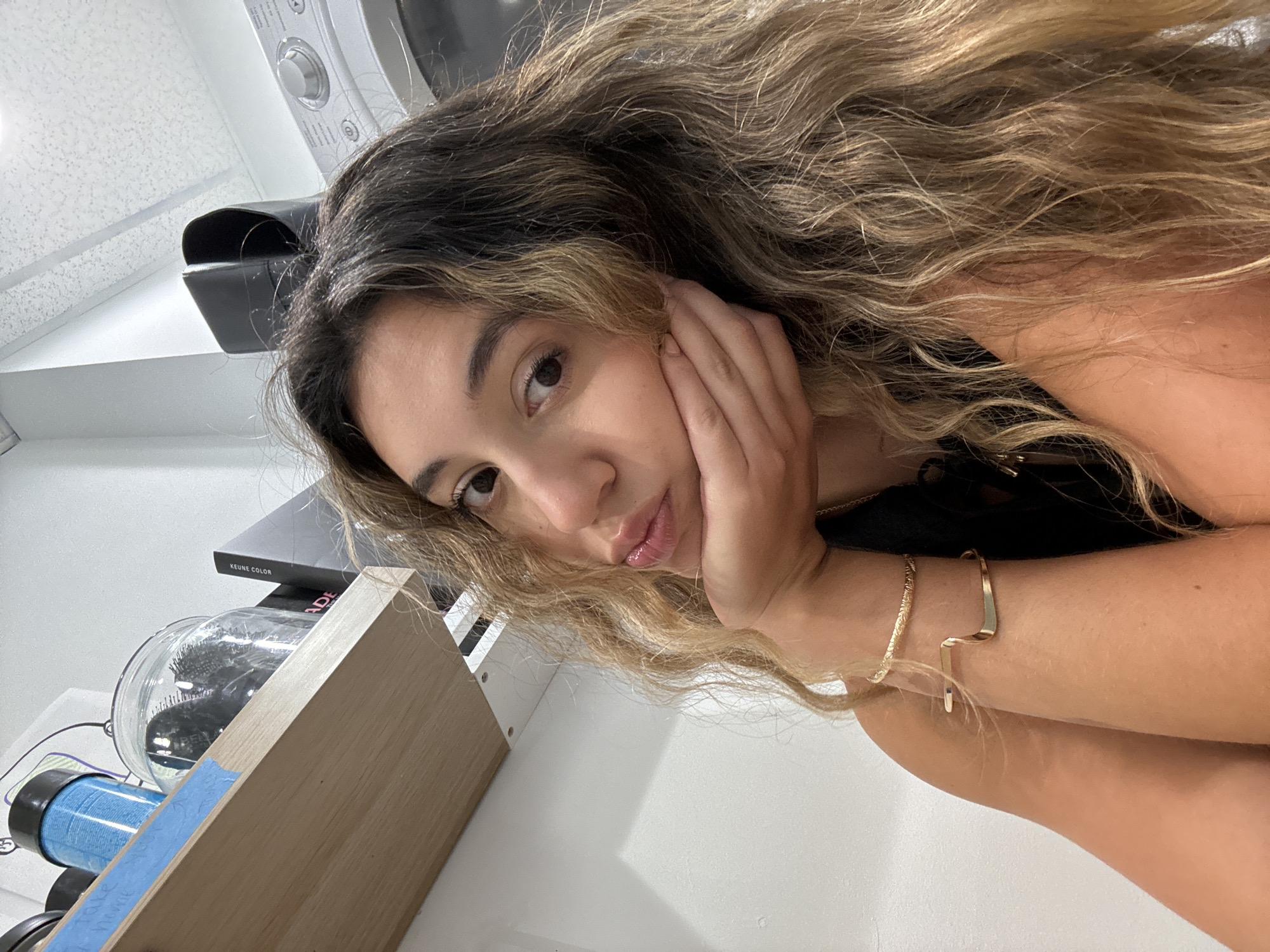 A young woman with long, wavy blonde hair rests her head on her hand, looking towards the camera. She is wearing a black top and a gold bracelet. In the background, a washing machine is visible, suggesting a laundry room or a home setting. To the left of the woman, a shelf holds several items, including a glass jar with hairbrushes inside, a blue hair roller, and a box labeled "KEUNE COLOR." The lighting in the image is bright and even, indicating it was likely taken indoors during the daytime. The woman's expression is soft and contemplative. The overall impression is that of a casual, personal moment captured in a domestic environment.