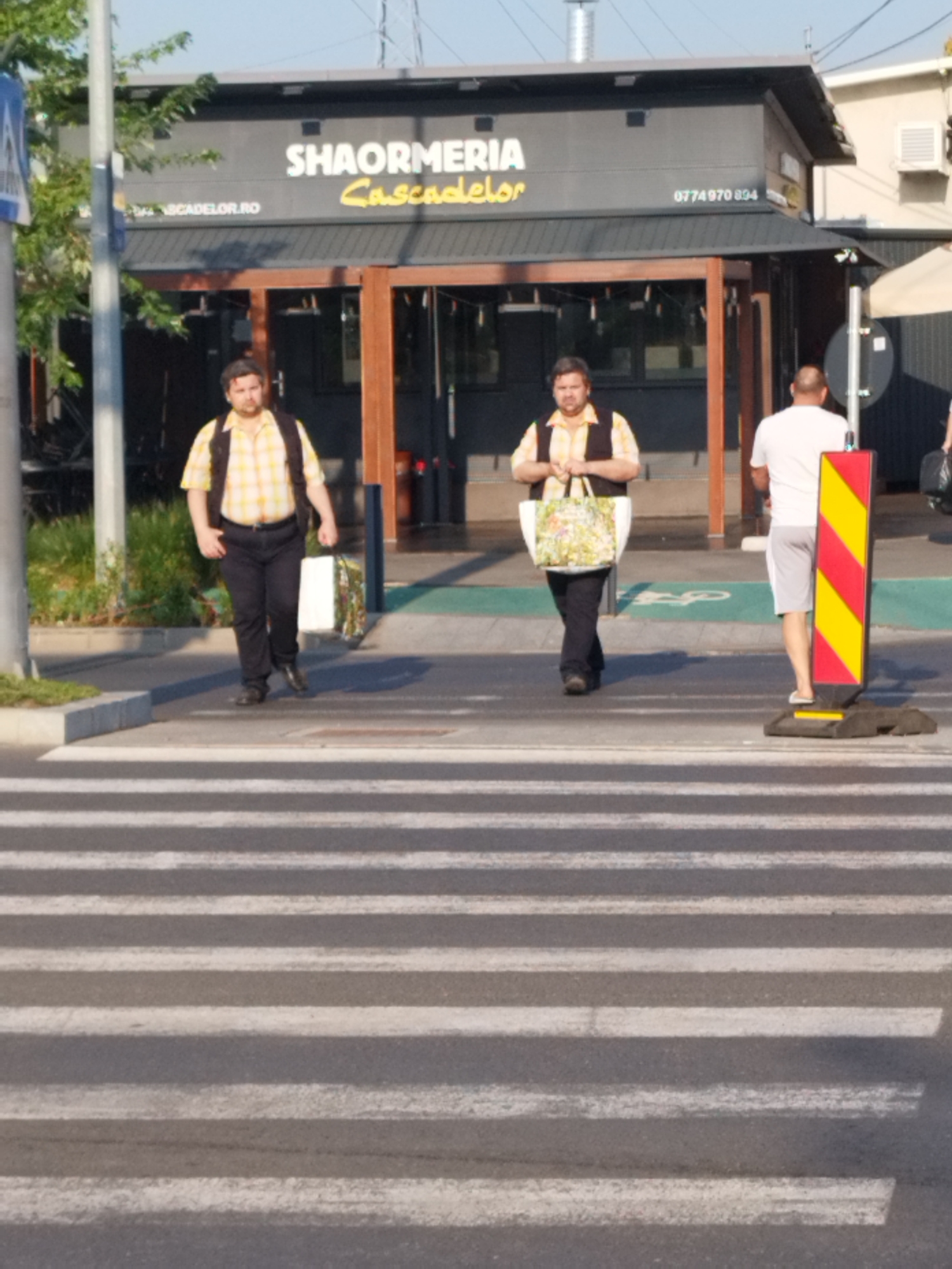Two men are walking across a pedestrian crossing in front of a restaurant in Bucharest, Romania. The restaurant, named "SHAORMERIA Cascadelor", has a sign with its name and a phone number. The man on the left is wearing a yellow plaid shirt and a dark vest over black pants, carrying a dark backpack. The man in the middle is wearing a similar yellow plaid shirt and dark vest with black pants, and is carrying a large patterned shopping bag. A third man, wearing a white t-shirt and light shorts, is standing on the sidewalk to the right of the pedestrian crossing, facing away from the camera. A yellow and red striped traffic cone is positioned in front of him. The environment appears to be an urban setting with a clear sky and some greenery in the background.