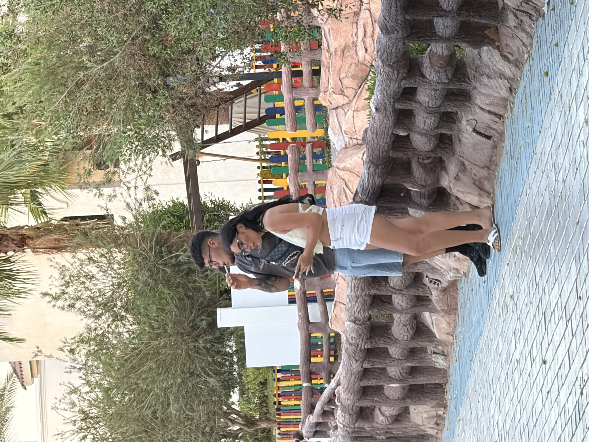 A young couple stands near a colorful fence in Sharm El Sheikh, Egypt. The man on the left, wearing glasses, a dark t-shirt, and light blue shorts, is holding a cigarette to his mouth. The woman beside him, also wearing glasses, a light yellow top, and white shorts, is looking at her phone. They are positioned on a paved walkway with a rustic wooden fence to their right, behind which is a playground with a ladder and swing set. The background shows trees and a building with red-tiled roofing.
