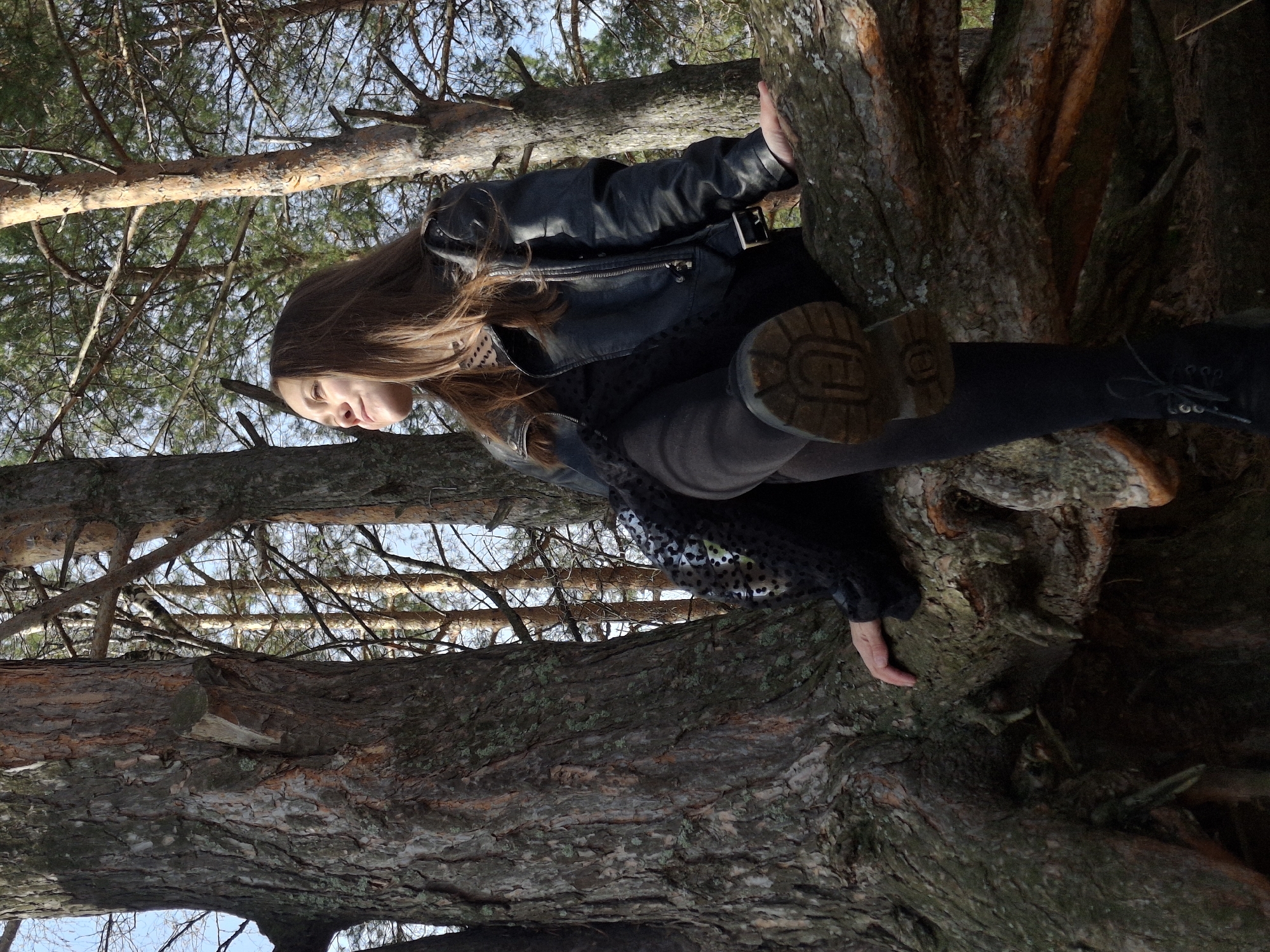A young woman with long brown hair is sitting in a tree. She is wearing a black leather jacket, a grey sweater, and black boots. Her right foot is extended towards the camera. The tree is in a forest, and the sky is visible through the branches. The location is likely in or near the city of Chaikovsky, Russia.