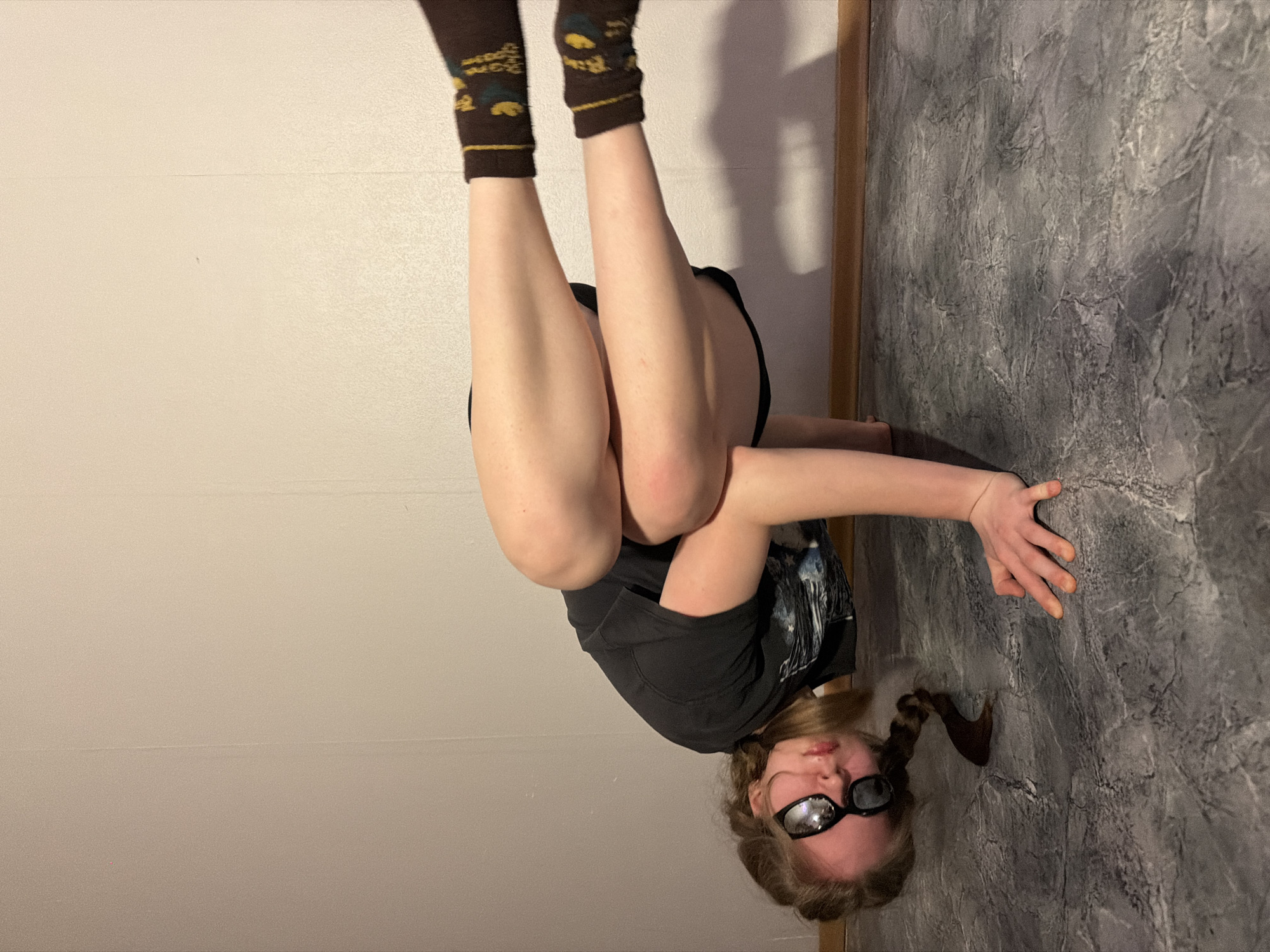 A person is positioned upside down in what appears to be an indoor setting. Their legs are elevated, with brown socks visible on their feet. The person's face is visible, and they are wearing sunglasses. One arm is extended towards a wall that has a gray, textured appearance, possibly resembling concrete or stone. A second wall is on the left which is white. The floor appears to be the surface the hand is touching, and there are no other people or subjects visible in the image. There are no indications in the image that suggest it was taken in Perm, Russia.
