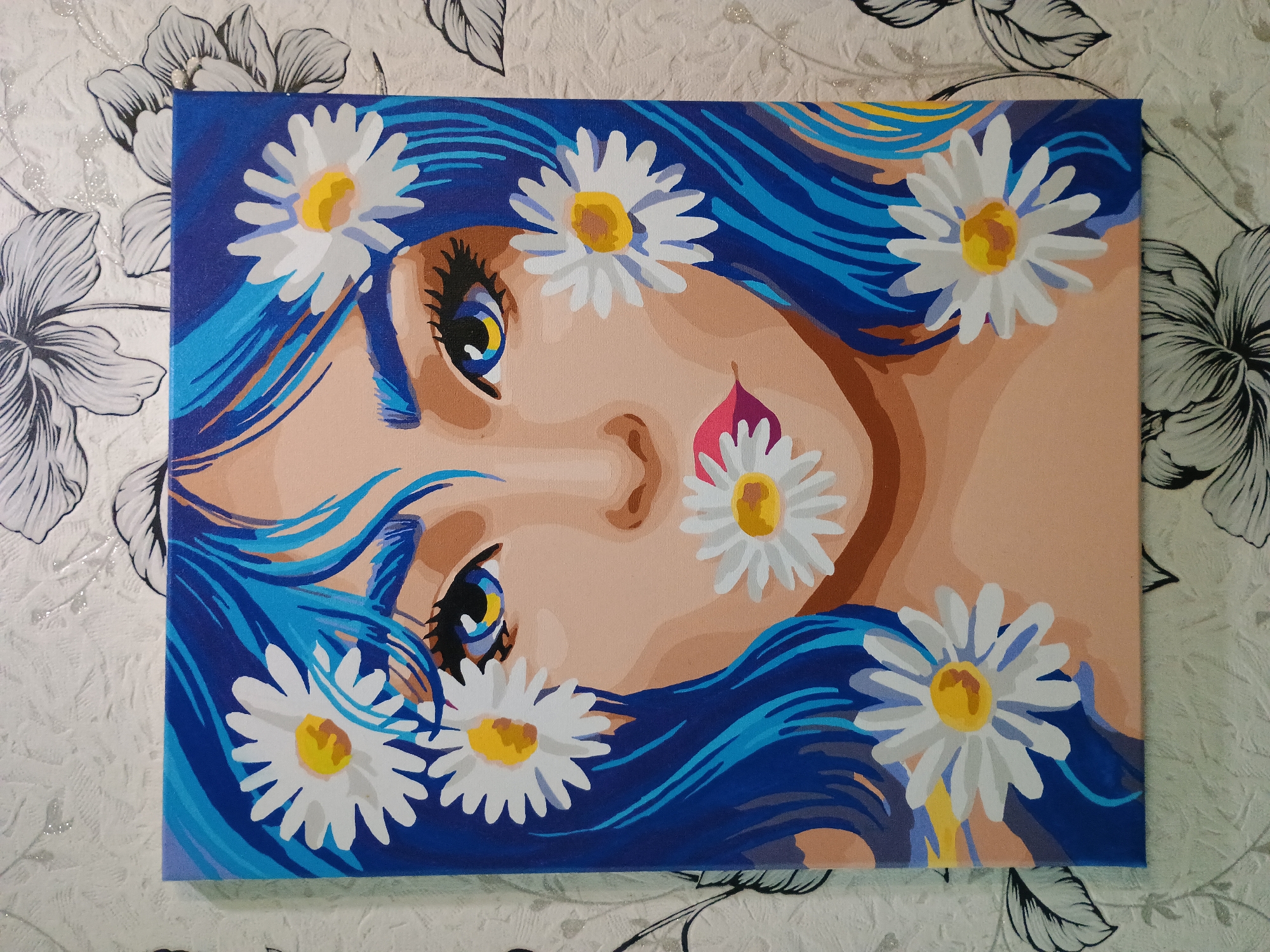 A painting on canvas depicts a stylized portrait of a young woman with vibrant blue hair. Her eyes are large and expressive with blue irises and yellow accents, framed by long, dark eyelashes. Daisies are artfully placed around her face, with one in her hair, one near her eye, one on her cheek, and one near her lips. Her lips are painted a deep pink with a hint of red. The background of the painting is a solid dark blue, contrasting with the woman's skin tone. The canvas is displayed on a wall covered with a floral-patterned wallpaper featuring a repeating design of black and white flowers. The overall impression is one of youthful beauty and a touch of whimsical artistry. There is no identifiable information within the image that would suggest a specific location in the city of Zaporizhzhia, Ukraine.