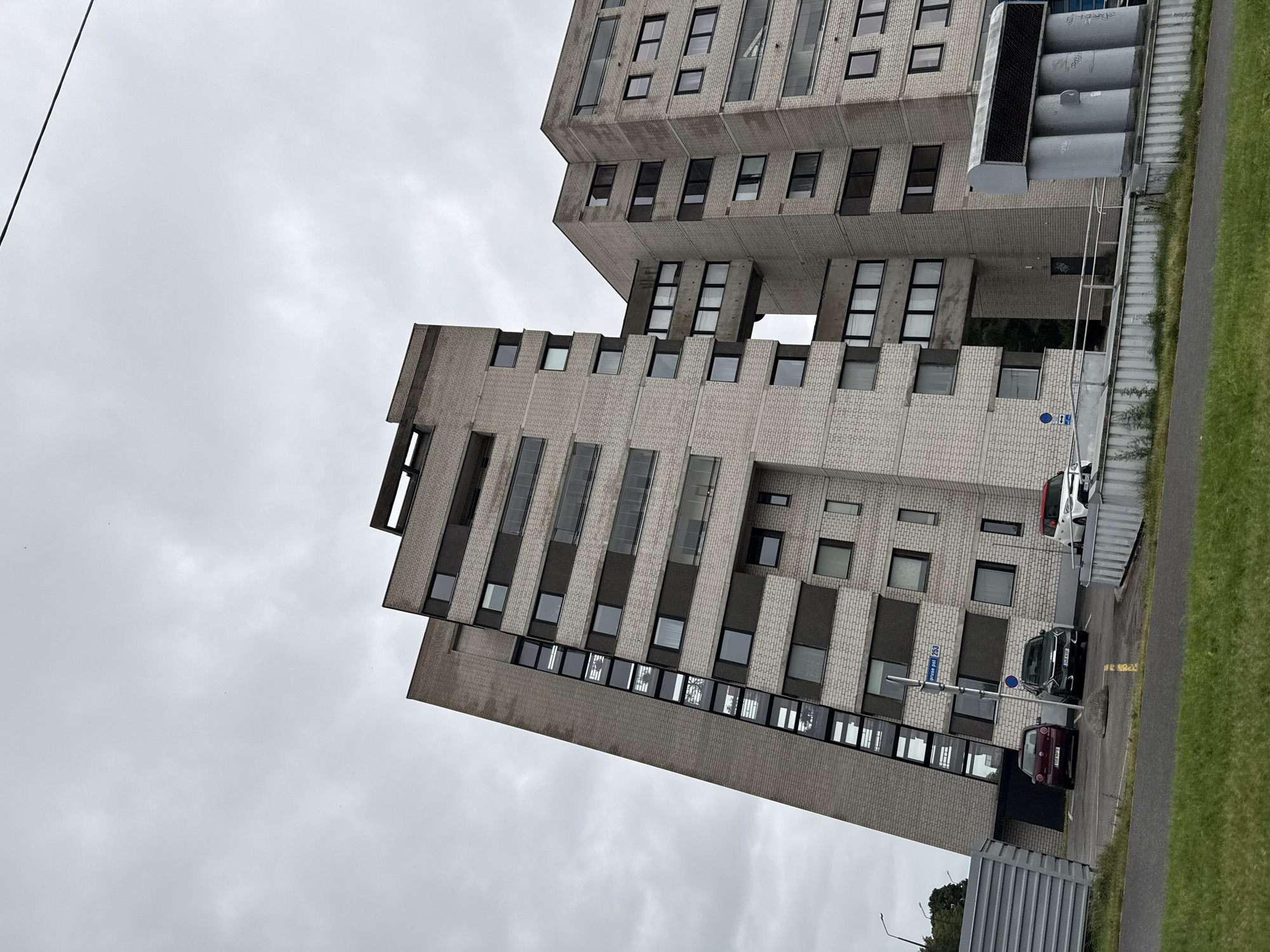 The image captures a view of a modern residential building with a unique architectural design in Tallinn, Estonia. The building features a light-colored brick facade with dark horizontal bands and numerous windows, some with balconies. One section of the building appears to be at a steep angle, creating a striking visual effect. In the foreground, there is a paved area with several parked cars, including a red Volkswagen Polo, a dark SUV, and a white and red car. A street sign reading "pruse tee 253" is visible. The environment appears overcast with a cloudy sky. The building's design suggests it is a multi-story apartment complex.