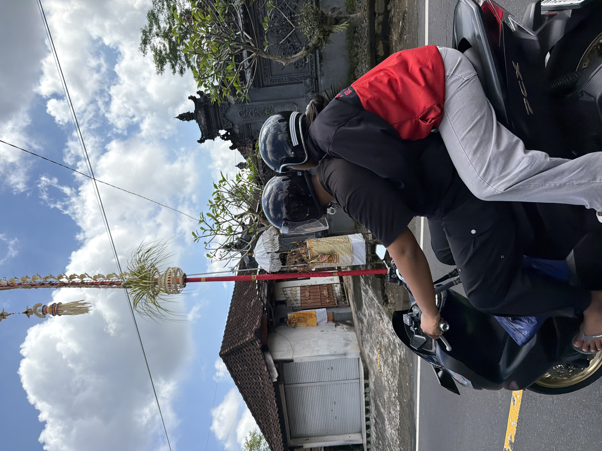 In Tampaksiring, Indonesia, two individuals are riding a black scooter on a paved road. The person in front is driving, wearing a black t-shirt and black pants, with a black helmet. The passenger behind them is wearing a red and black jacket and light grey pants, also with a black helmet, and is embracing the driver. The background shows a traditional Balinese building with a tiled roof and a decorative gate, as well as a tall red pole adorned with offerings and greenery. The sky is partly cloudy.