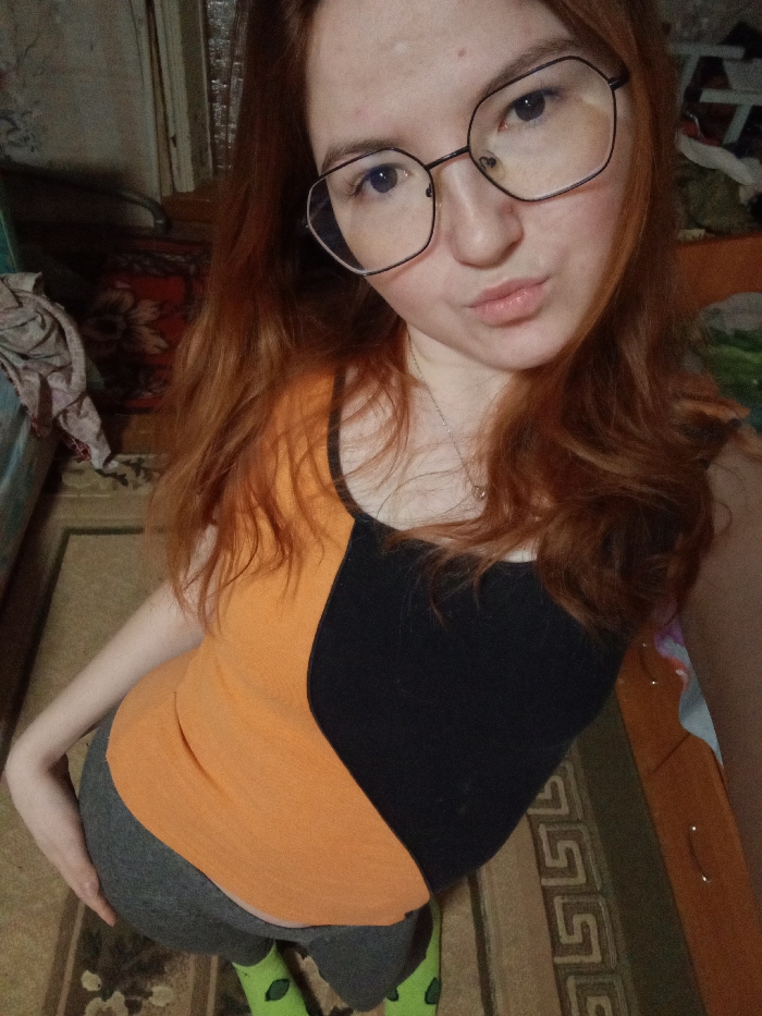 An indoor, high-angle portrait depicts a young female subject with long, reddish-brown hair, light-colored eyes, and facial blemishes, wearing dark-rimmed, hexagonal eyeglasses. She exhibits a neutral expression with slightly pouted lips and a visible silver chain with a small pendant around her neck. The subject is dressed in a sleeveless top, horizontally split into an orange upper section and a black lower section, grey form-fitting leggings, and bright green socks adorned with a repetitive pattern. Her left hand is positioned on her left hip or upper buttock. The background indicates a residential room in Старый Шагирт, Russia, featuring a patterned floor rug with geometric designs in beige, brown, and green tones. A dark red tapestry with a floral motif is mounted on a light-colored wall to the left, adjacent to a partially visible light blue furniture item draped with crumpled fabric. A wooden multi-drawer dresser is partially visible on the right side of the frame. The scene is illuminated by what appears to be ambient indoor lighting.