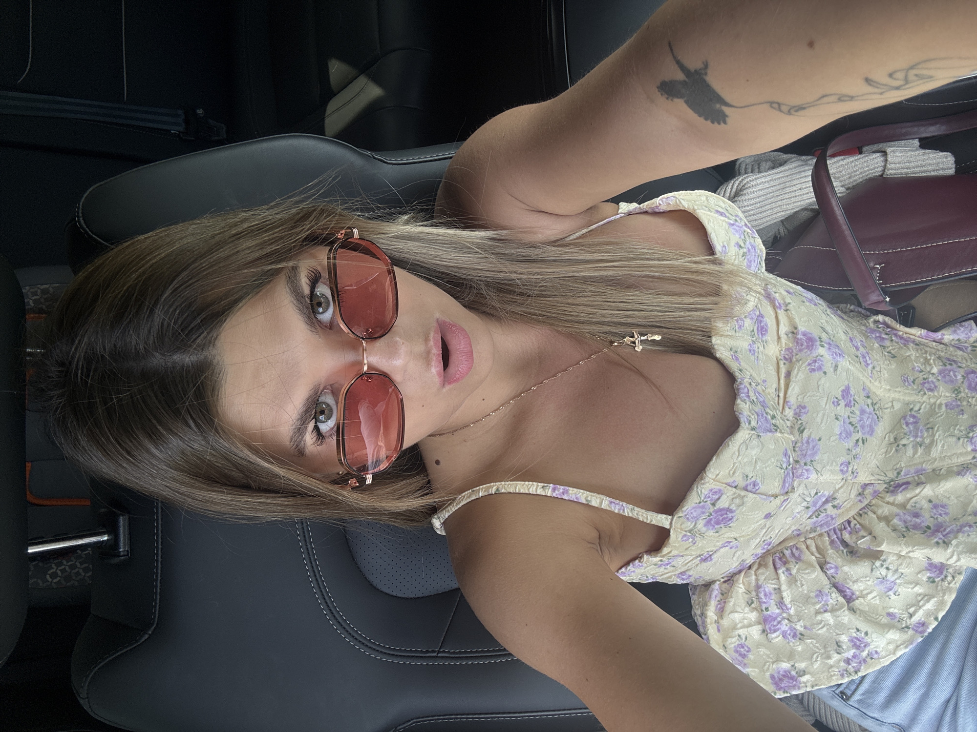 A young woman with blonde hair and sunglasses is taking a selfie in a car. She is wearing a yellow floral corset top and blue jeans. Her mouth is slightly open, and she is wearing a necklace with a cross pendant. There is a tattoo on her arm. The background shows the interior of a car, with black leather seats. The location is identified as a street in the city of Pastukhovo, Russia.