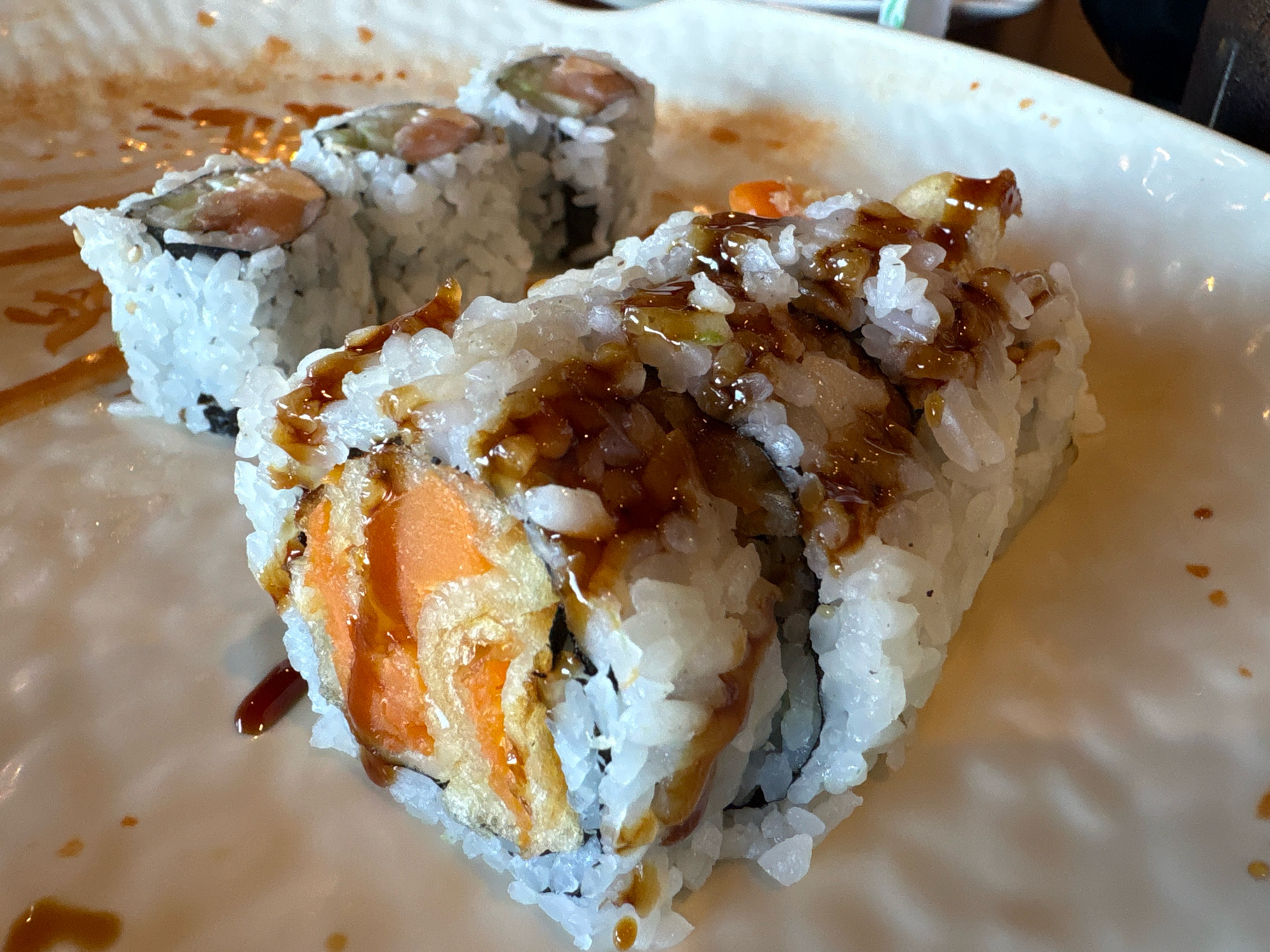 The image shows a close-up, overhead view of sushi rolls on a white plate. The sushi is covered in a dark sauce and appears to be partially eaten. The background is out of focus, but suggests a restaurant setting. The image was taken in Rocky Hill, Connecticut, United States of America.