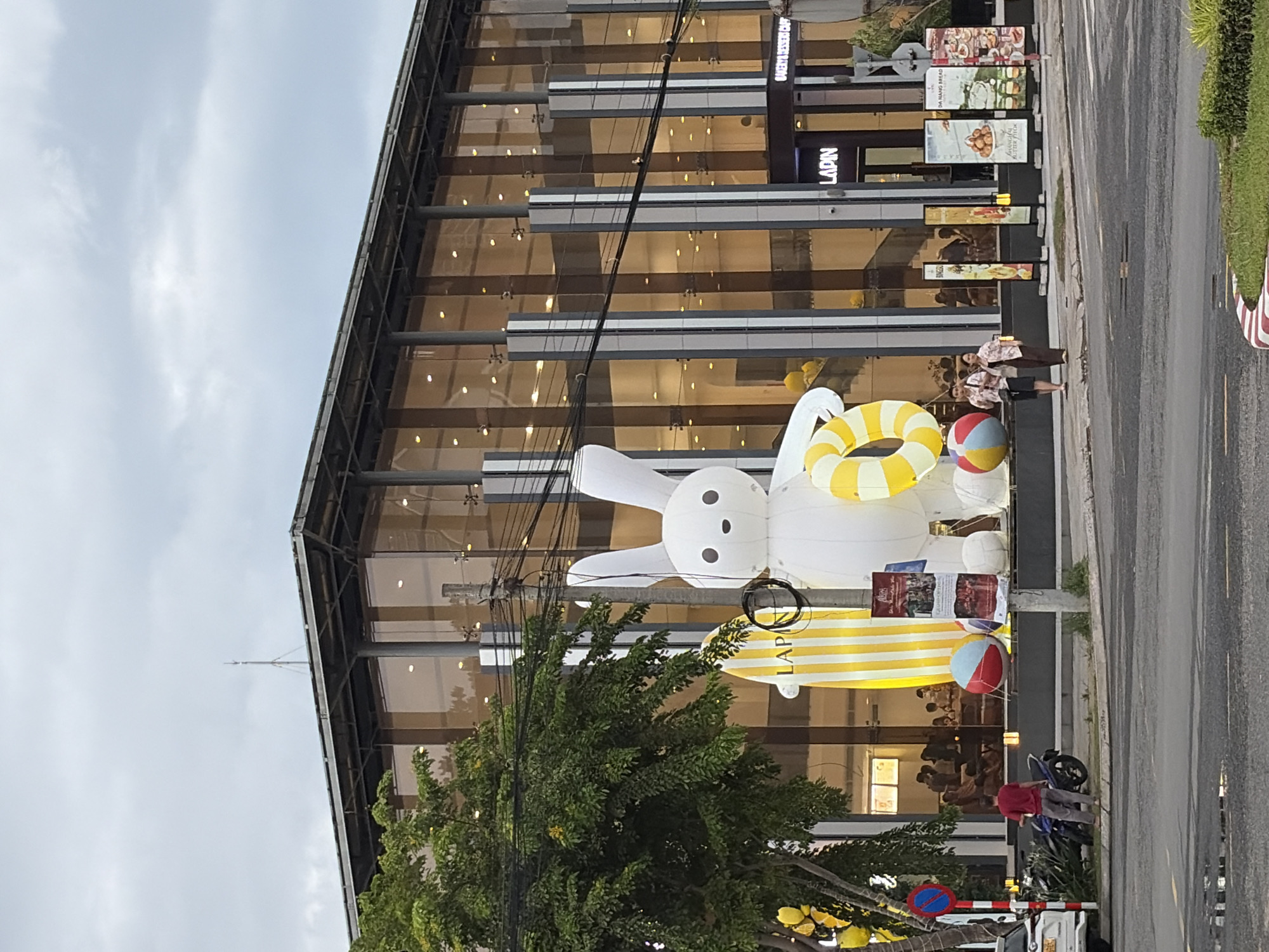 The image shows a street view in Da Nang, Vietnam, with a modern building featuring a glass facade. In front of the building, there is a large inflatable white bunny holding a yellow and white striped surfboard and a yellow and white ring. Several beach balls are placed around the bunny. A street sign with a red circle and a blue diagonal line is visible on the left. A white car is parked on the left side of the road, and a motorcycle is parked near the building. People are visible inside the building, and two women are walking on the sidewalk in front of the building. The building has signs that say "LAPIN" and "BAKERY & DESSERT CAFE". The overall scene appears to be a commercial area in Da Nang.