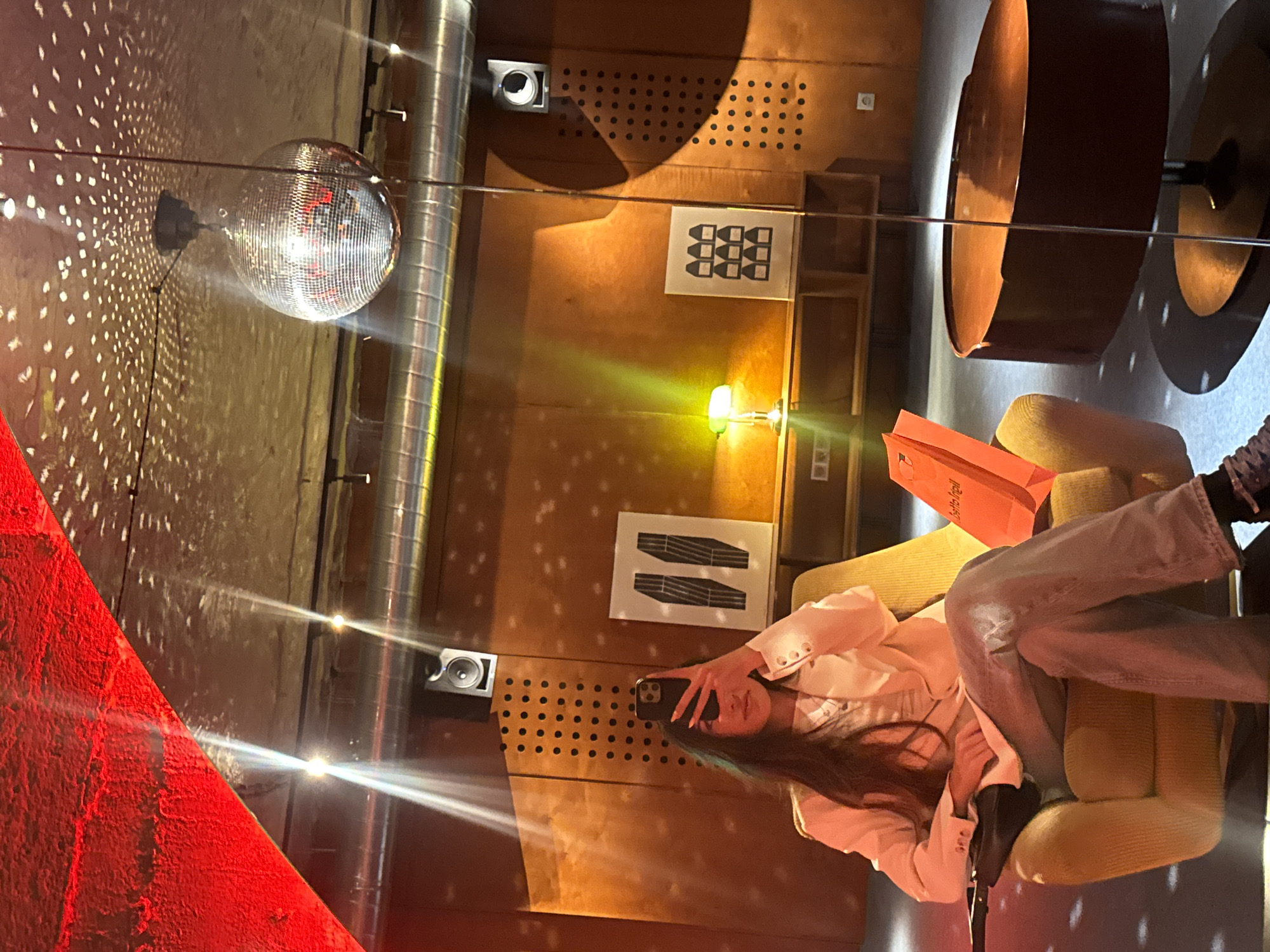 A woman is sitting in a yellow chair, holding a smartphone, in a room illuminated by a disco ball. The location is Yerevan, Armenia. The room has a copper-colored wall adorned with patterned decals and mounted speakers. A red object is visible on the lower left side. She is wearing a white jacket and light jeans. An orange shopping bag is situated next to her on the chair.