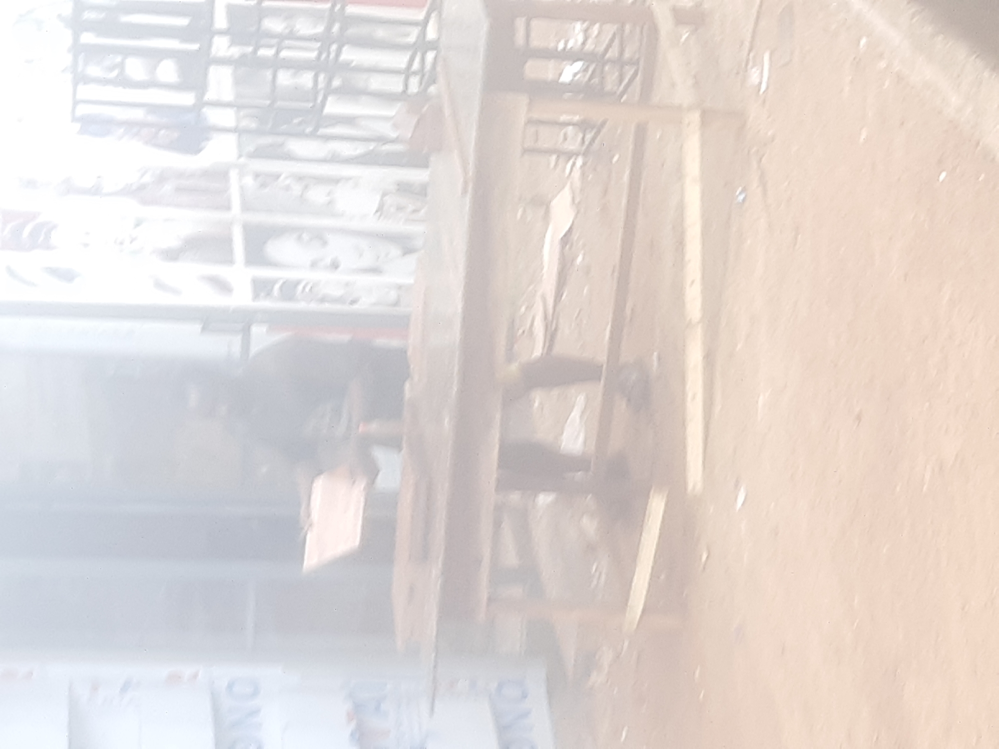 The image captures an outdoor scene in Enugu, Nigeria, characterized by a dusty, sandy ground and a general haze that softens details. In the mid-ground, a person, appearing to be male, stands behind a wooden table. The person is blurred but seems to be engaged in an activity involving the table. The table itself is a rustic wooden structure with a visible top and legs, set on the uneven ground. To the left of the table, a portion of a structure with lettering is visible, likely a building or a sign, with the partial word "NO" discernible. In the background, more structures and possibly some caged displays or shelves are present, also obscured by the atmospheric haze and low image quality. The overall impression is of a marketplace or a street-side stall.