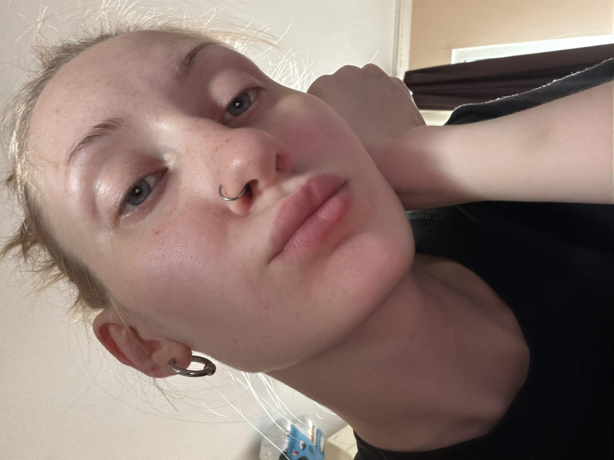 The image is a close-up, tilted selfie of a young woman with blonde hair. She is wearing a black top and has a silver hoop earring in her left ear and a silver septum piercing. Her right arm is bent and resting near her face, with her hand touching her cheek. Her eyes are a light blue, and her lips are slightly parted. She has a natural makeup look, with a few freckles visible on her nose and cheeks. The background appears to be indoors, with a wall and a curtain visible. The lighting suggests it could be daytime. There is no visible text in the image. The context provided suggests the location is Ladozhskoe Ozero, Russia.