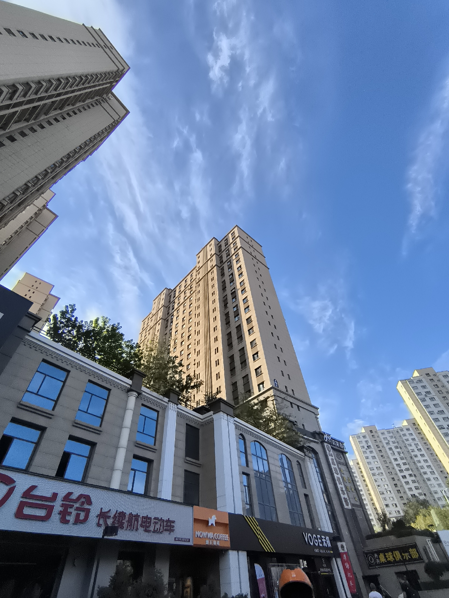 The image is an upward-angle shot of several tall buildings in a city under a bright blue sky with wispy clouds. In the foreground, a low-rise building with grey facade and blue-tinted windows is visible. A red Chinese sign is prominently displayed on this building, with Chinese characters "台铃 长续航电动车" (Tailing Long-Endurance Electric Vehicle). To the right of this sign, a smaller orange sign reads "NOWWA COFFEE". Above these commercial establishments, greenery from trees is visible.
Dominating the mid-ground and background are several high-rise residential or office buildings. One tall, beige building directly in the center features numerous windows and a textured facade, appearing to be lit by sunlight. To the left, another, narrower tall building of similar color and design is angled against the sky. Further to the right, another cluster of multi-story buildings, also with a grid of windows, forms the backdrop. The architecture suggests a modern urban environment.
The image's perspective looking upwards emphasizes the scale of the architecture against the expansive sky. No people are clearly discernible in the main body of the image, though silhouettes might be present at the ground level. The overall scene depicts a daytime urban landscape. Based on visual cues such as the specific Chinese signage, the style of the buildings, and the context of the request specifying Ürümqi, China, this scene is located within the city of Ürümqi.