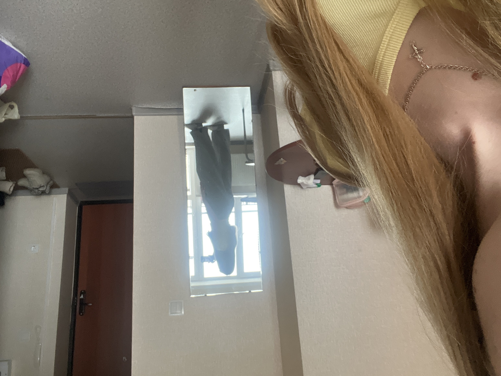 The image captures an indoor scene, likely a room, with a person taking a selfie. In the immediate foreground, the upper right portion of a person's body is visible, featuring long, light-colored hair flowing downwards. The person wears a light yellow top, and a silver chain necklace with a small cross charm adorns their neck and collarbone area.
Centrally placed on a light-colored wall is a rectangular mirror, reflecting the lower half of a person (presumably the same individual taking the photo) wearing dark pants. Beyond the reflected person, a brightly lit window with a grid of panes is visible in the mirror, indicating daytime.
The room features light-colored, possibly beige or off-white, walls and a light gray ceiling. To the left, a dark brown door with a black frame and handle is partially visible, next to some light-colored cabinetry or shelving. On a surface to the right of the mirror, a few small objects, including a reddish-pink item and some crumpled white material, are present.
The overall activity is a self-portrait being taken in a casual, personal space. There is no visible text or specific location cues within the image itself.
