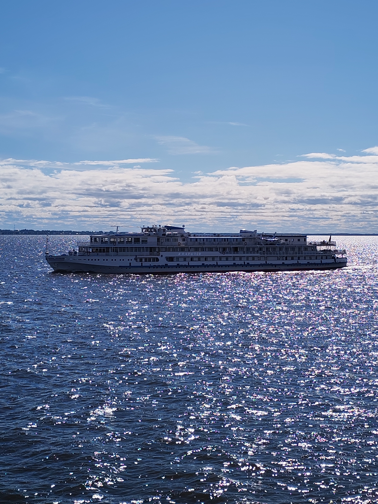 The image captures a multi-deck passenger cruise ship on a body of water under a partly cloudy sky. The water surface is highly reflective, sparkling in the sunlight. The landscape in the distance is a low strip on the horizon, indicating a lakeshore or riverbank. The scene is in the city of Belozersk, Russia.