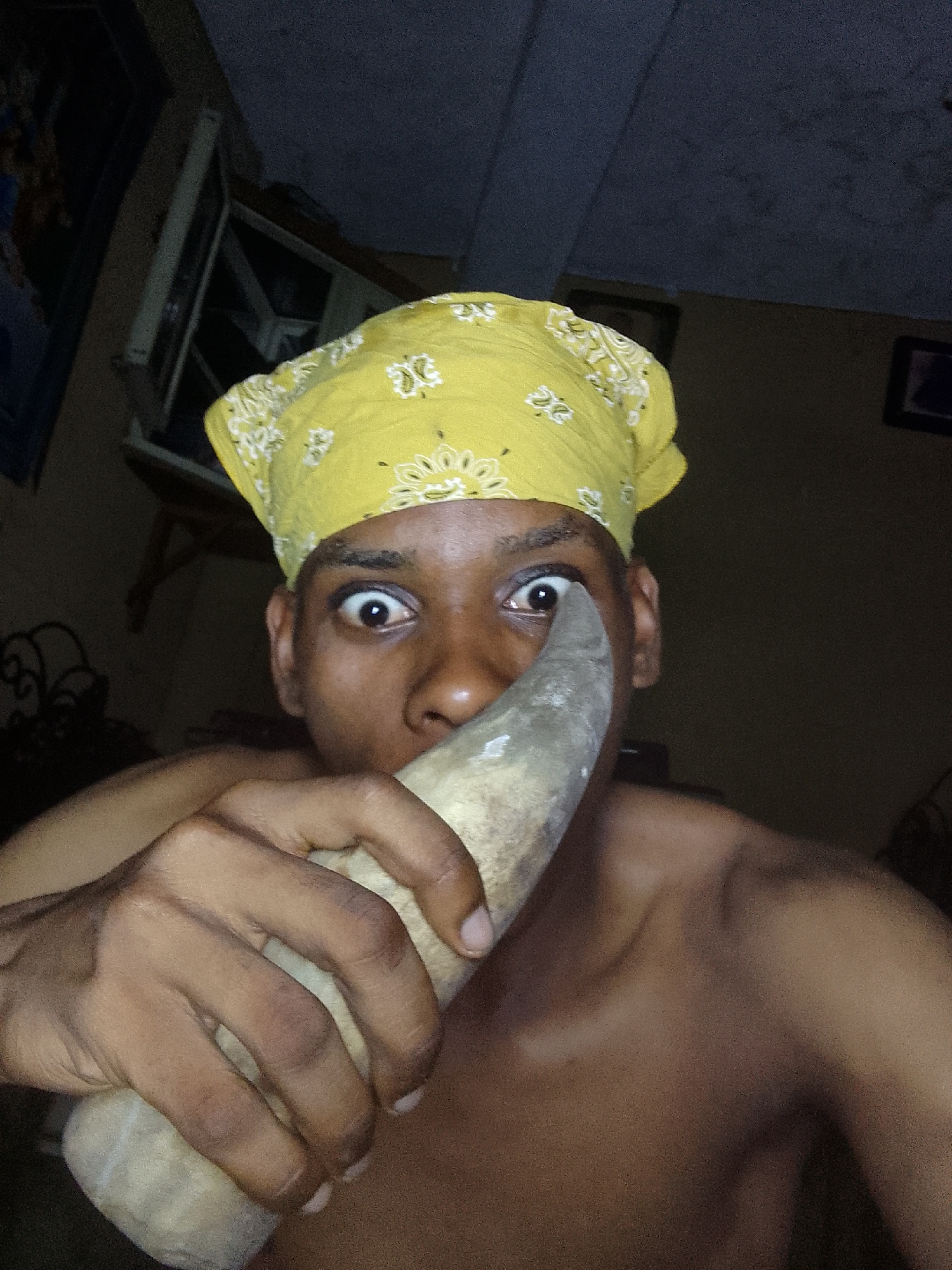 A close-up shot depicts a shirtless individual with dark skin and a yellow bandana on their head. The person is holding a gray object, possibly a horn, close to their face, partially obscuring their mouth. Their eyes are wide and expressive. The background is dark and indistinct, showing a partially visible window frame, a piece of furniture, and a framed picture on the wall. The location is not discernible from the provided image.