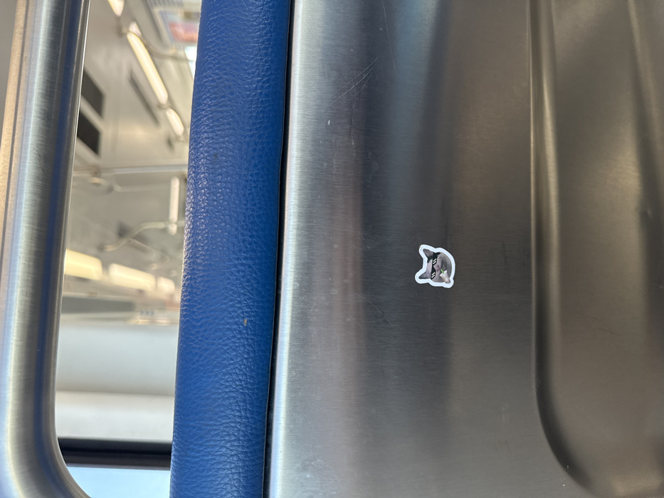 The image displays the interior of a public transit vehicle, identified as a Metrorail train car, situated within Hialeah, United States of America. In the immediate foreground, a segment of a royal blue, textured vinyl cushion, likely part of a seat or barrier, is visible. Below this cushion is a brushed metallic panel. Centered on this metallic surface, slightly to the left, is a small, die-cut sticker, approximately 3 centimeters in width. The sticker depicts the head of a gray cat wearing black, pixelated sunglasses and holding a small, white, cylindrical object in its mouth. The metallic panel exhibits minor scuff marks. The background, rendered out of focus, reveals the train car's ceiling, overhead fluorescent strip lighting, and multiple metallic grab rails and straps, consistent with public transportation infrastructure. No individuals are present or discernable.