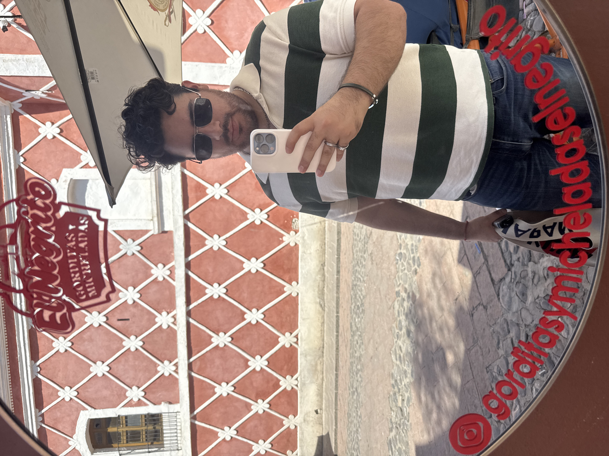 A man is taking a selfie in a round mirror at "El Negrito Gorditas y Micheladas" in Bernal, Mexico. He is wearing a green and white striped shirt, jeans, sunglasses, and jewelry (bracelet, rings, necklace). He holds a white phone with a triple camera lens. The mirror reflects the cobblestone street, the building facade with a distinctive red and white diamond pattern, and an umbrella. The restaurant's name and social media handle "gorditassymichelbernal" are written in red around the mirror's edge.