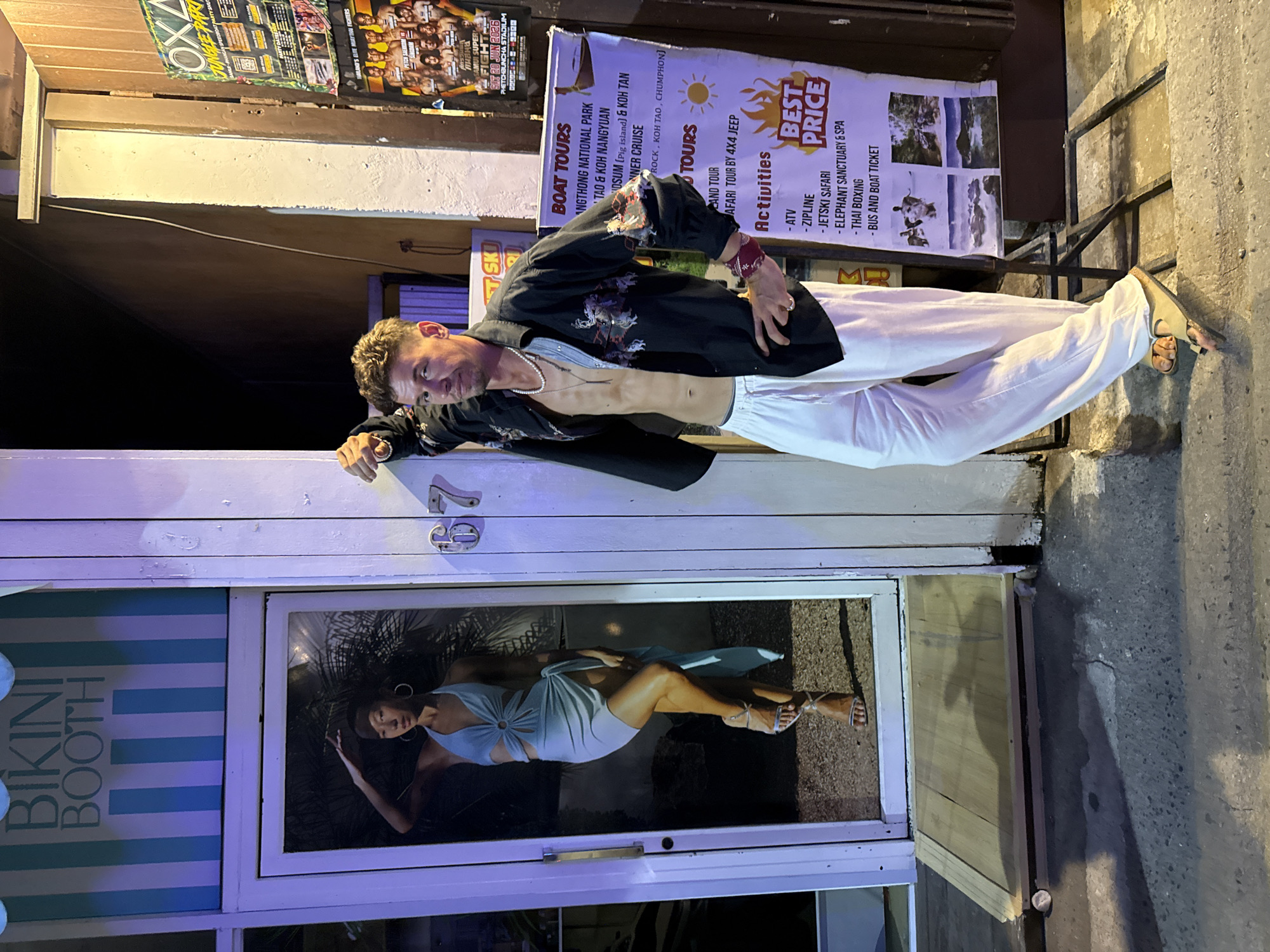A man stands outside a "BIKINI BOOTH" with a poster advertising boat tours and activities. He is wearing a black, open-chested shirt with an embroidered pattern, white linen pants, and tan sandals. A pearl necklace and a red bracelet are visible. The man is leaning against a white pillar with the number "67" attached. Inside the booth's glass door, a full-length poster of a woman in a light blue cutout dress and silver heels is displayed. The background is dark, suggesting nighttime. A ramp made of wooden planks is placed on the ground leading to the entrance. The location is identified as Baan Bo Phut, Thailand, as indicated by the boat tour poster.