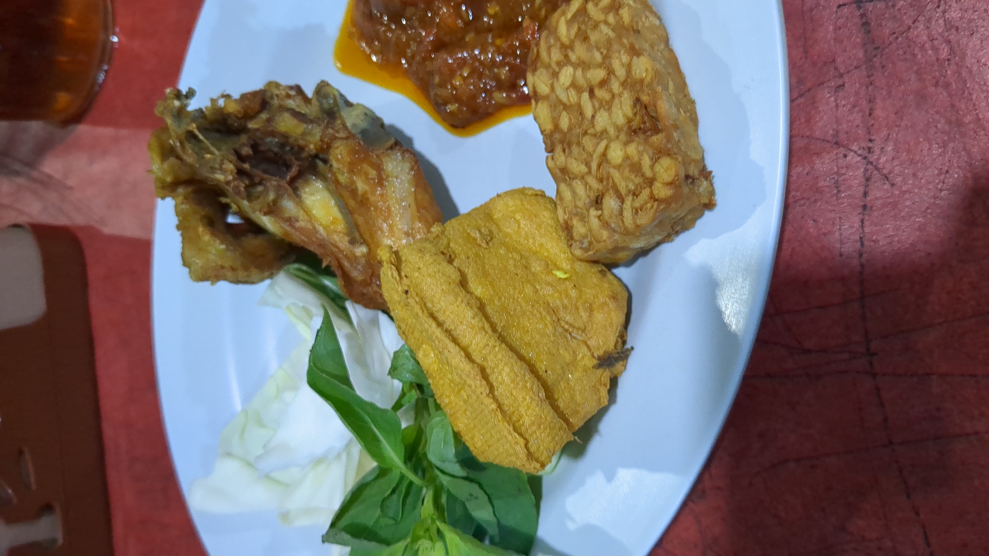 A close-up shot captures a plate of Indonesian food, likely served at a casual dining establishment in Purwakarta, Indonesia. The plate, white and round, is filled with several fried items: a piece of fried chicken, a large piece of fried tofu, and a smaller rectangular piece of fried tempeh. A dollop of reddish sambal sauce is present to the right of the chicken. Fresh green basil leaves and a few pieces of shredded white cabbage garnish the left side of the plate. The food is presented on a red textured surface, possibly a tablecloth or table. The lighting suggests an indoor setting, potentially a restaurant or warung.