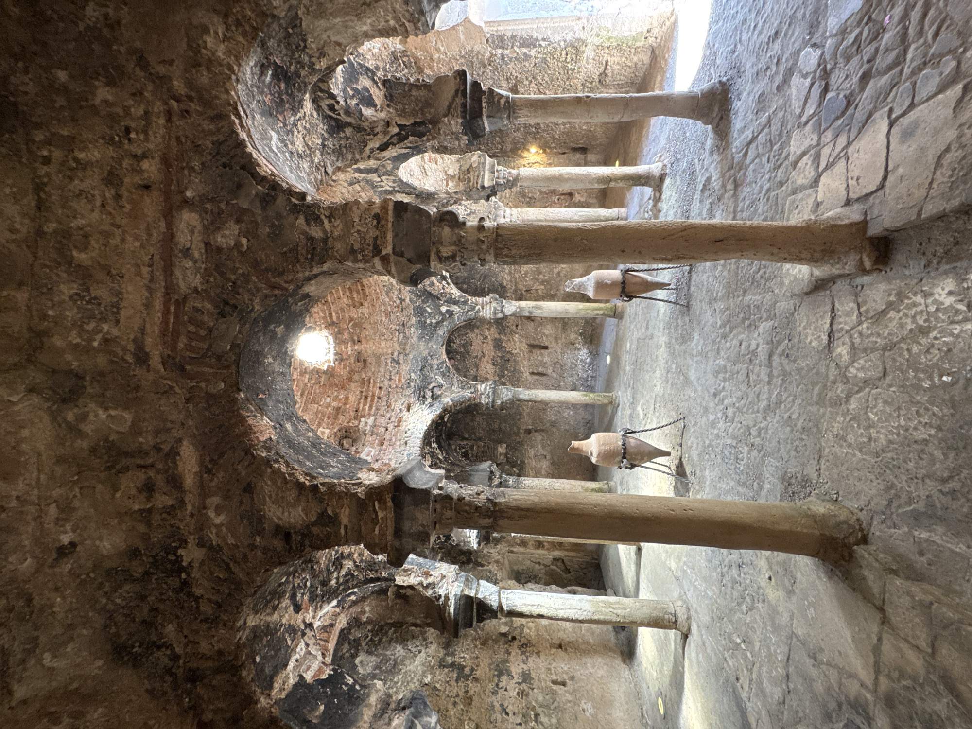 This is a wide-angle, eye-level photograph of the interior of the historic Arab Baths (Baños Árabes) located in Palma de Mallorca, Spain. The scene depicts a vaulted chamber with rough, textured stone walls and ceiling, exhibiting signs of age and wear. Several stone columns, some with intricately carved capitals, support the arched ceiling. Light filters in from a circular opening in the ceiling and from an opening on the right side of the frame. Two large, antique clay amphorae, each supported by a metal stand, are placed on the stone-paved floor. The floor itself is uneven and composed of various sizes of stone slabs. The overall atmosphere is one of historical preservation and antiquity.