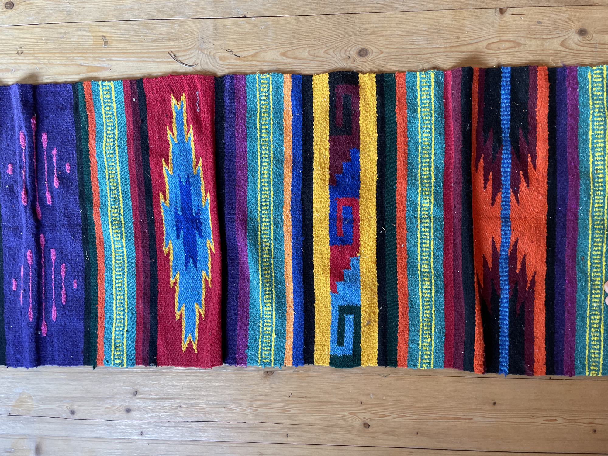 The image shows a woven textile laid out horizontally on a wooden floor. The textile is a long, narrow runner with a vibrant, striped pattern featuring various colors including purple, pink, green, orange, yellow, blue, black, and red. There are geometric designs woven into the fabric, such as diamond shapes and zigzags. Some sections have thin, repetitive yellow lines running vertically. On the left side, there is a purple section with pinkish-purple vertical dashes. The right side of the textile shows a repeating pattern of orange, red, and black stripes with a blue flame-like motif in the center. The wooden floor appears to be made of light-colored planks with visible grain and some small knots. The lighting suggests it is daytime, and there are no obvious clues about the specific location or any activity taking place. No text is visible in the image.
