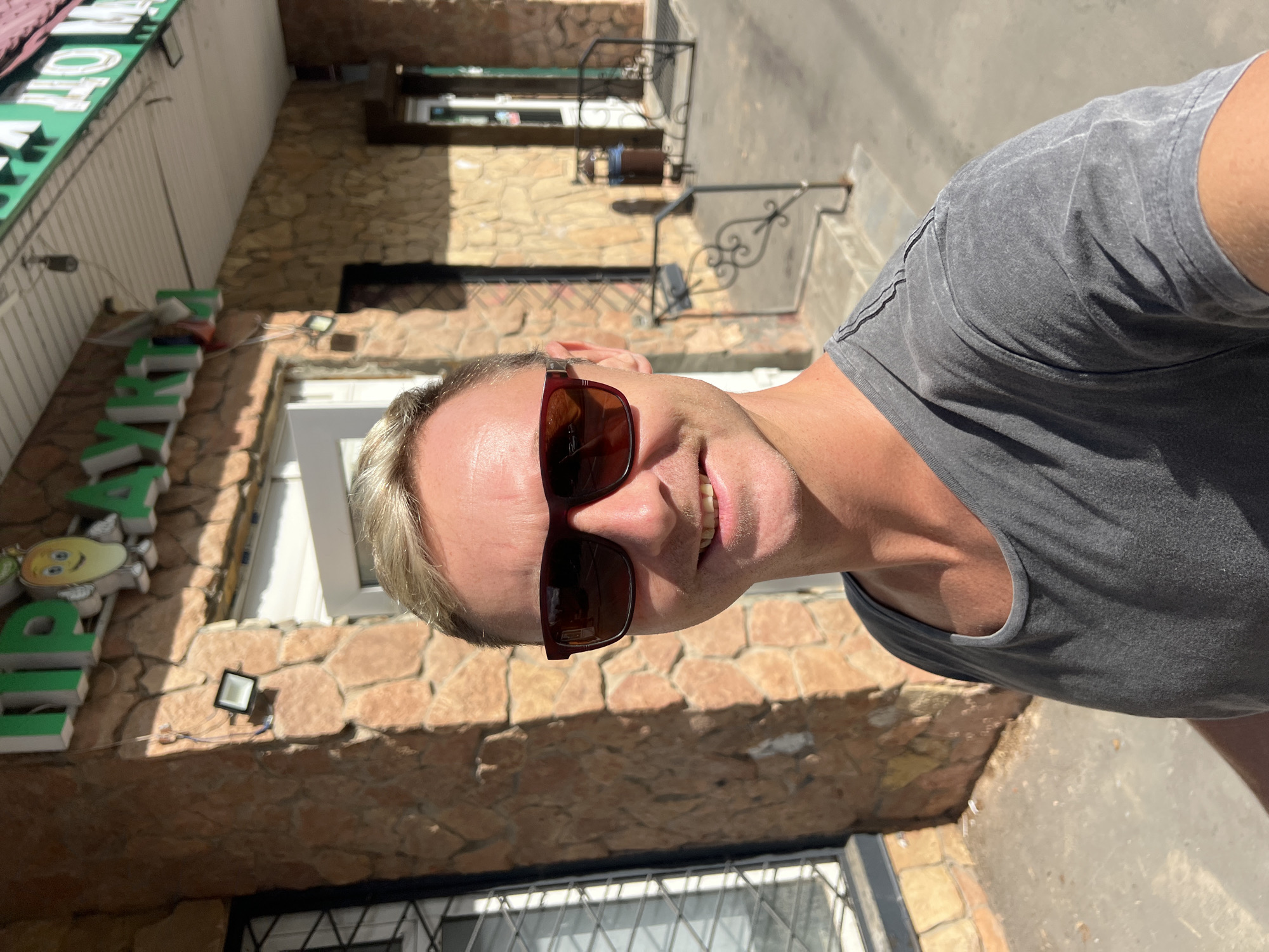 A man with blond hair and sunglasses takes a selfie in front of a stone building with a green sign. The sign has text in Russian and an illustration of a cartoon mango. The building appears to be a shop or market. The man is wearing a gray t-shirt. The ground is paved, and there are some railings and a trash can in the background. The lighting suggests it is daytime. The location is identified as Shishkin Les, Russia.