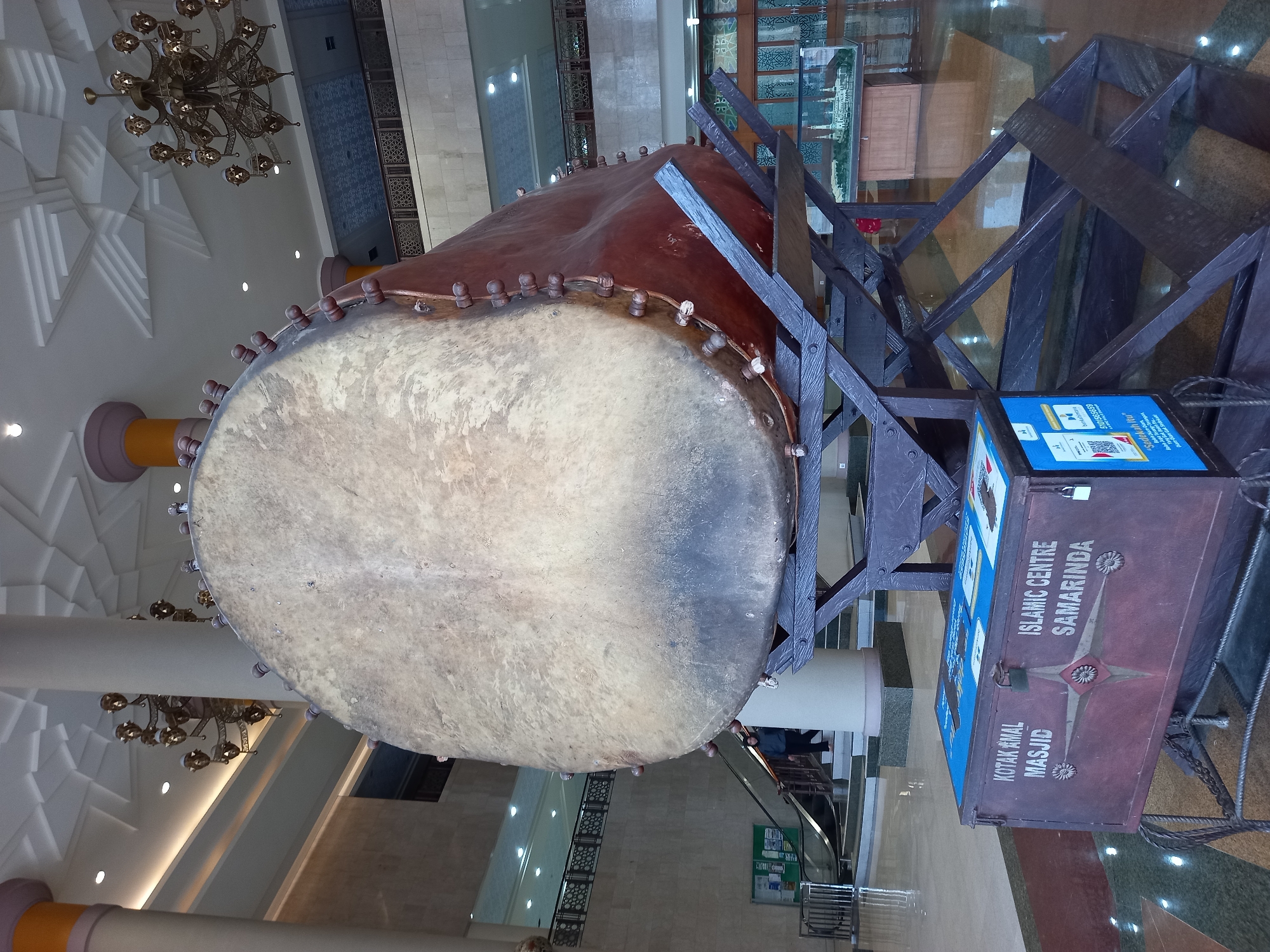 A large, weathered drum is displayed on a dark wooden stand inside a building. The drum is positioned centrally, dominating the frame, with its head facing the viewer. It appears to be made of hide stretched over a wooden frame and is adorned with numerous rounded pegs along its circumference. In the foreground, a decorative donation box with blue and maroon coloring sits on a metal frame. The box has text in Indonesian, including "KOTAK AMAL MASJID" and "ISLAMIC CENTRE SAMARINDA," indicating its purpose and connection to the Islamic Centre in Samarinda, Indonesia. The interior space features marble flooring, tall columns, ornate chandeliers, and modern architectural elements, suggesting it is a public building such as a lobby or convention center. In the background, an escalator and a person descending it can be seen, further confirming the indoor setting.