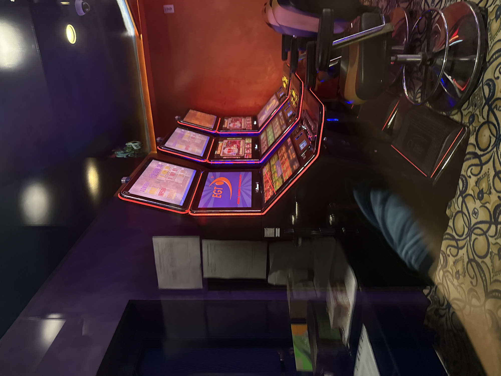 The image depicts a casino interior in Pleven, Bulgaria. Multiple electronic slot machines are arranged in a curved row, each with illuminated screens displaying game interfaces. A spinning stool with chrome detailing stands in the foreground. Adjacent to the machines is a counter surface cluttered with papers and personal items. The overall lighting in the casino appears dim and atmospheric.