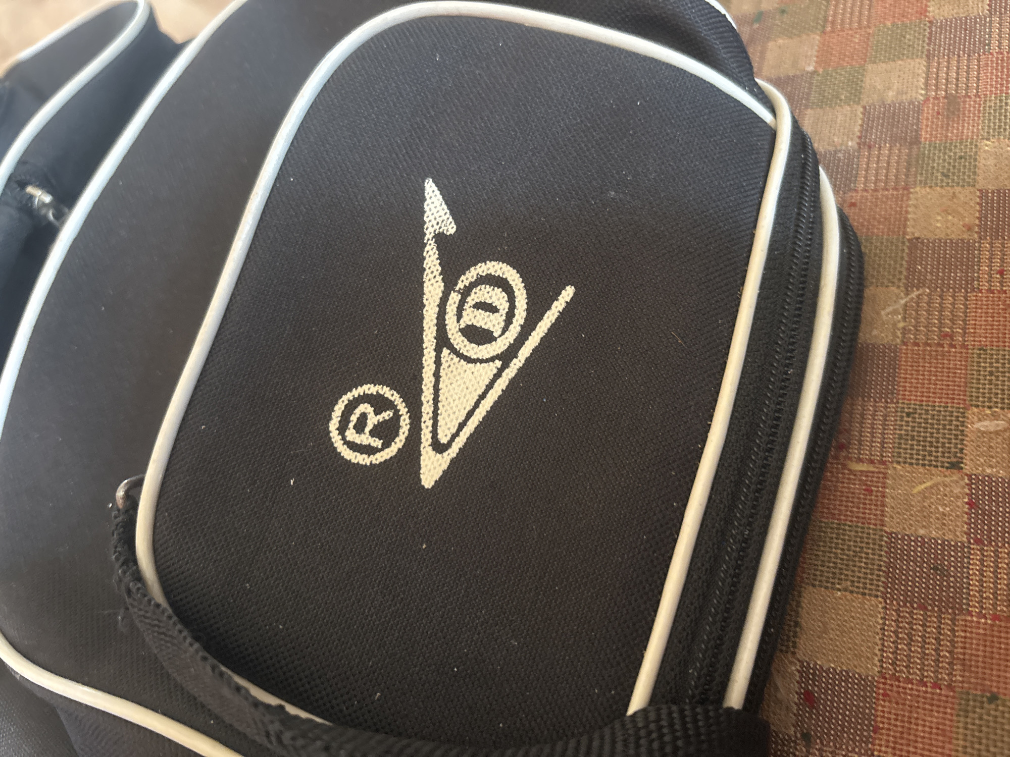 The image displays a close-up of a black textile bag with white piping, placed on a surface with a brown, tan, and reddish checkered pattern. The bag is positioned diagonally, with its top-left corner extending out of view and the bottom-right corner also partially cropped.
The central focus is a white logo printed on the front flap of the black bag. The logo features a prominent upward-pointing triangular or arrow-like shape. To the left of this main design is a clear registered trademark symbol (®). Within the main logo, a circular element encloses a stylized character that resembles a lowercase 'e' or a similar letter. The white lines of the logo are slightly textured, suggesting a printed or embroidered application on the fabric.
Visible details of the bag include a zipper running along its right side, outlined by the white piping, indicating a compartment. A black fabric strap or handle is also partially visible at the bottom. The checkered surface beneath the bag has a woven texture. The lighting suggests an indoor setting or a shaded outdoor area in Asenovgrad, Bulgaria. No people or specific activities are depicted.