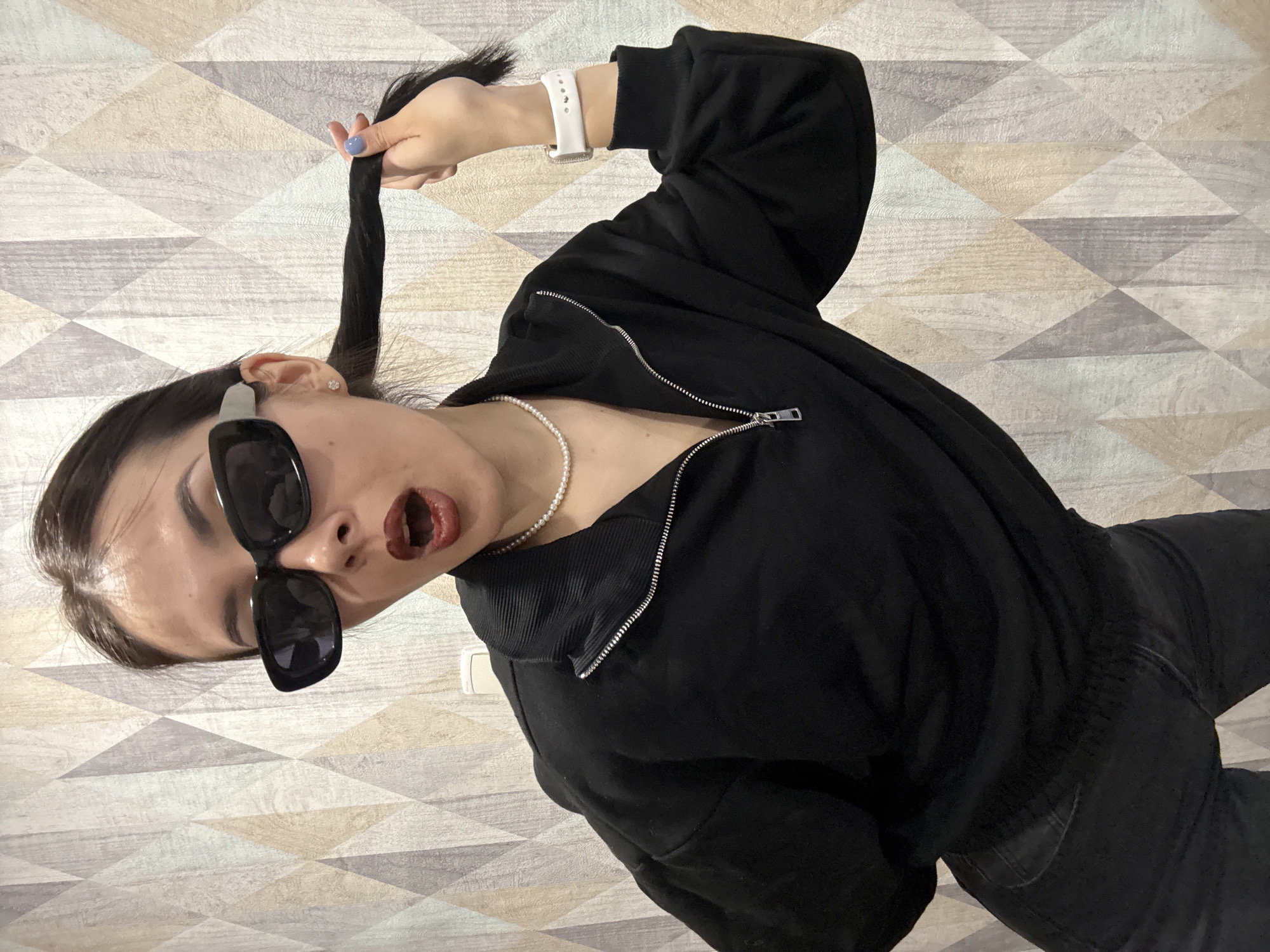 A young woman with dark hair pulled into a high ponytail, wearing oversized sunglasses, a pearl necklace, and a black zippered jacket, is posing in an indoor setting. She is holding her ponytail up with her right hand, which has blue-colored nails, and wearing a white smartwatch. Her expression appears to be dramatic, with her mouth open. She is wearing dark lipstick. She is wearing dark jeans. The background is a wall covered in a geometrically patterned wallpaper. There is no information available to determine the location is in Votkinsk, Russia.