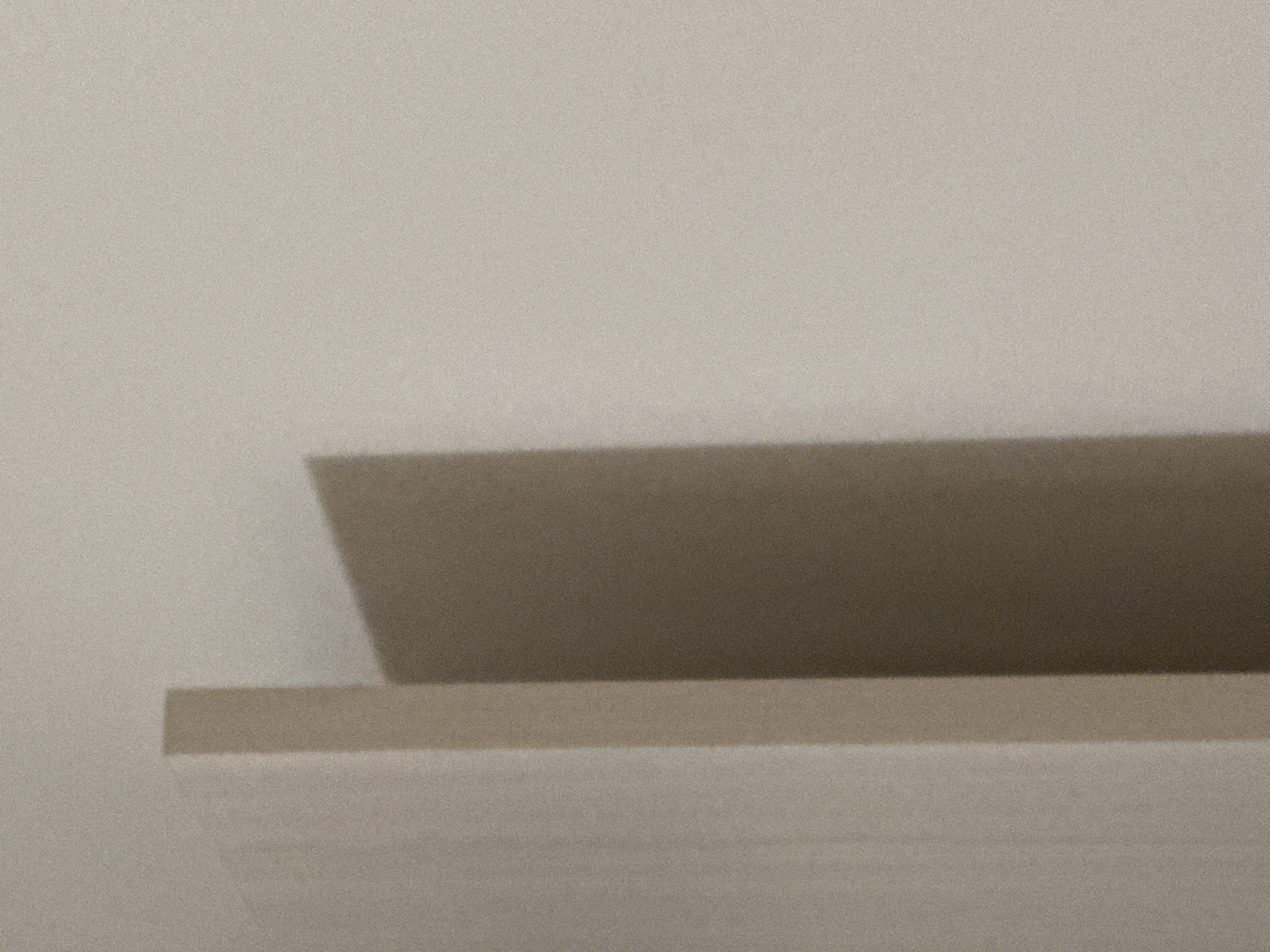 The image depicts a close-up, low-angle shot of a corner where two walls meet, casting a shadow. The lighting is soft, creating a subtle gradient across the surfaces. The texture of the wall appears slightly grainy, consistent with a painted or textured finish. There are no people, animals, or distinct objects visible; the focus is solely on the architectural elements and the play of light and shadow. The scene is abstract and minimalist. Without additional context or identifiable landmarks, it is impossible to determine the specific location within Budva, Montenegro.
